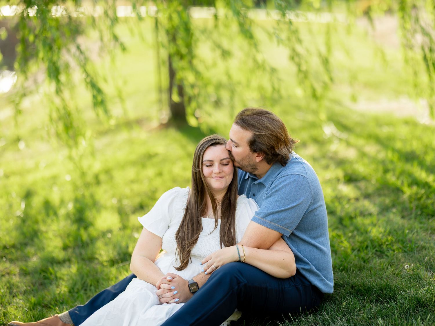 Couple sitting on grass under willow tree, man kissing woman's cheek during outdoor engagement photo session.