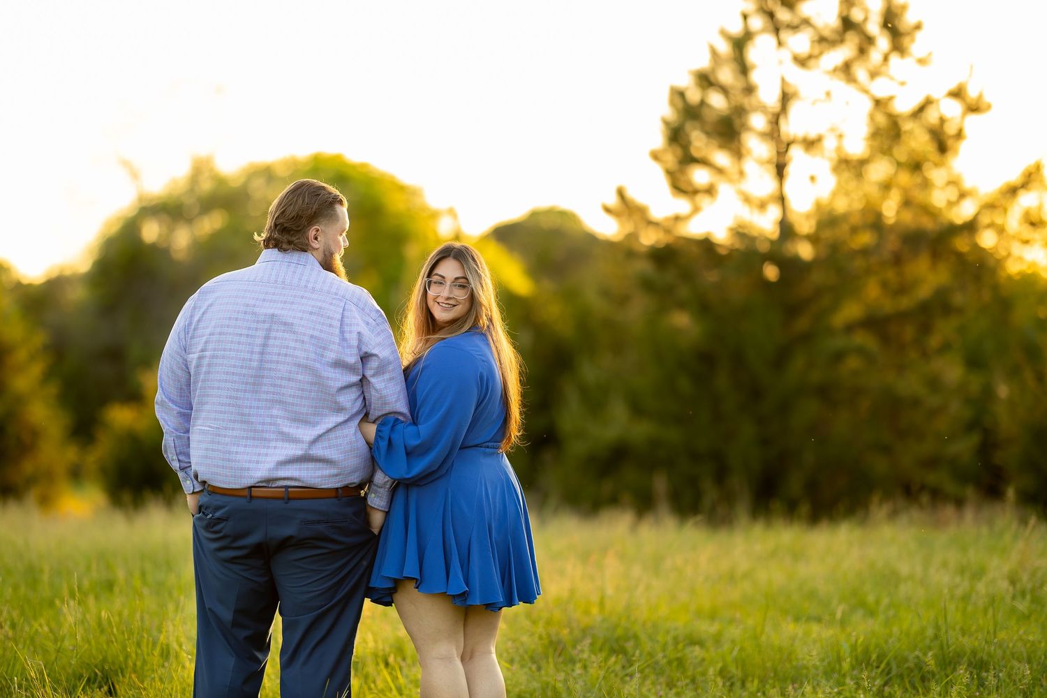 Couple posing at golden hour in a green field, woman in blue dress embracing man in striped shirt.
