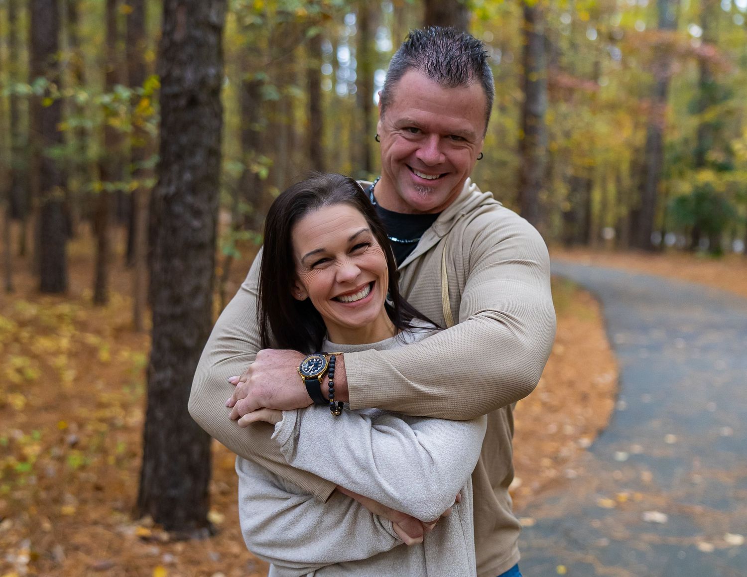 A couple embraces while wearing matching beige sweaters on an autumn path surrounded by fall foliage.