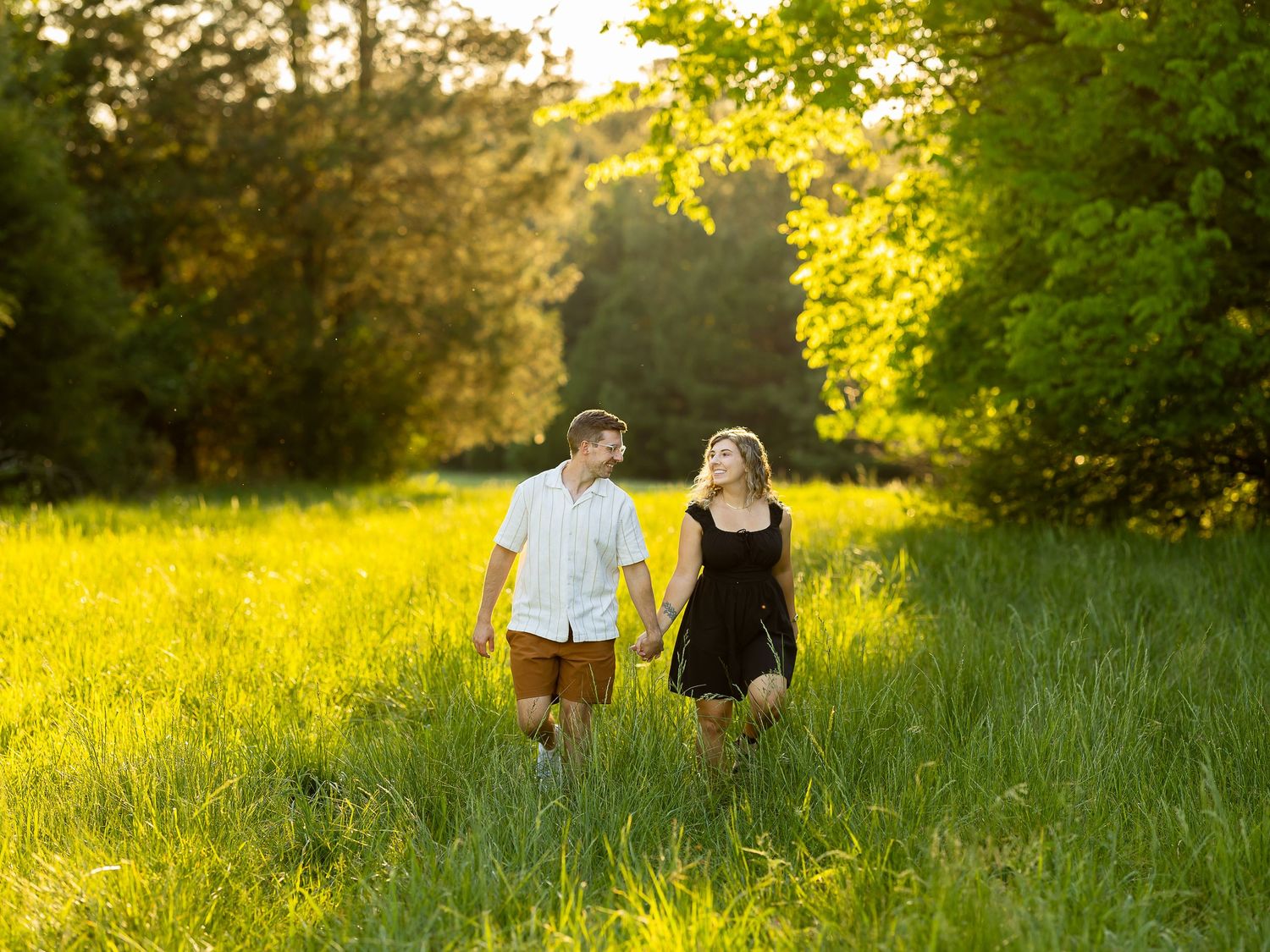 Couple holding hands walking through lush green meadow bathed in golden sunset light surrounded by trees.
