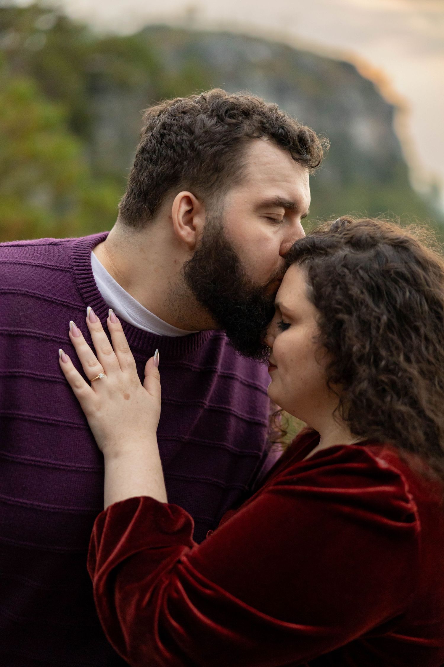 A romantic couple shares an intimate moment outdoors wearing burgundy and purple sweaters against a blurred natural background.