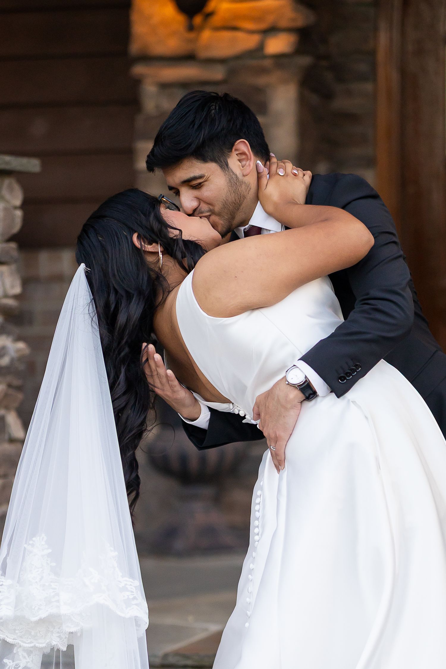 Groom dipping and kissing bride in white gown and veil outside rustic wooden venue.
