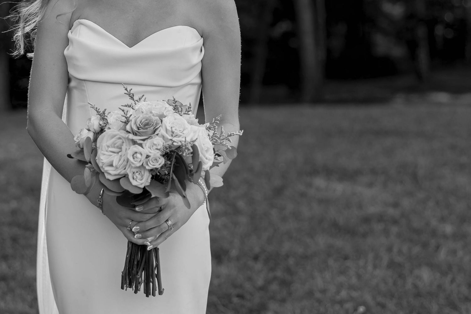 Black and white photo of a bride in a strapless gown holding a rose bouquet outdoors.