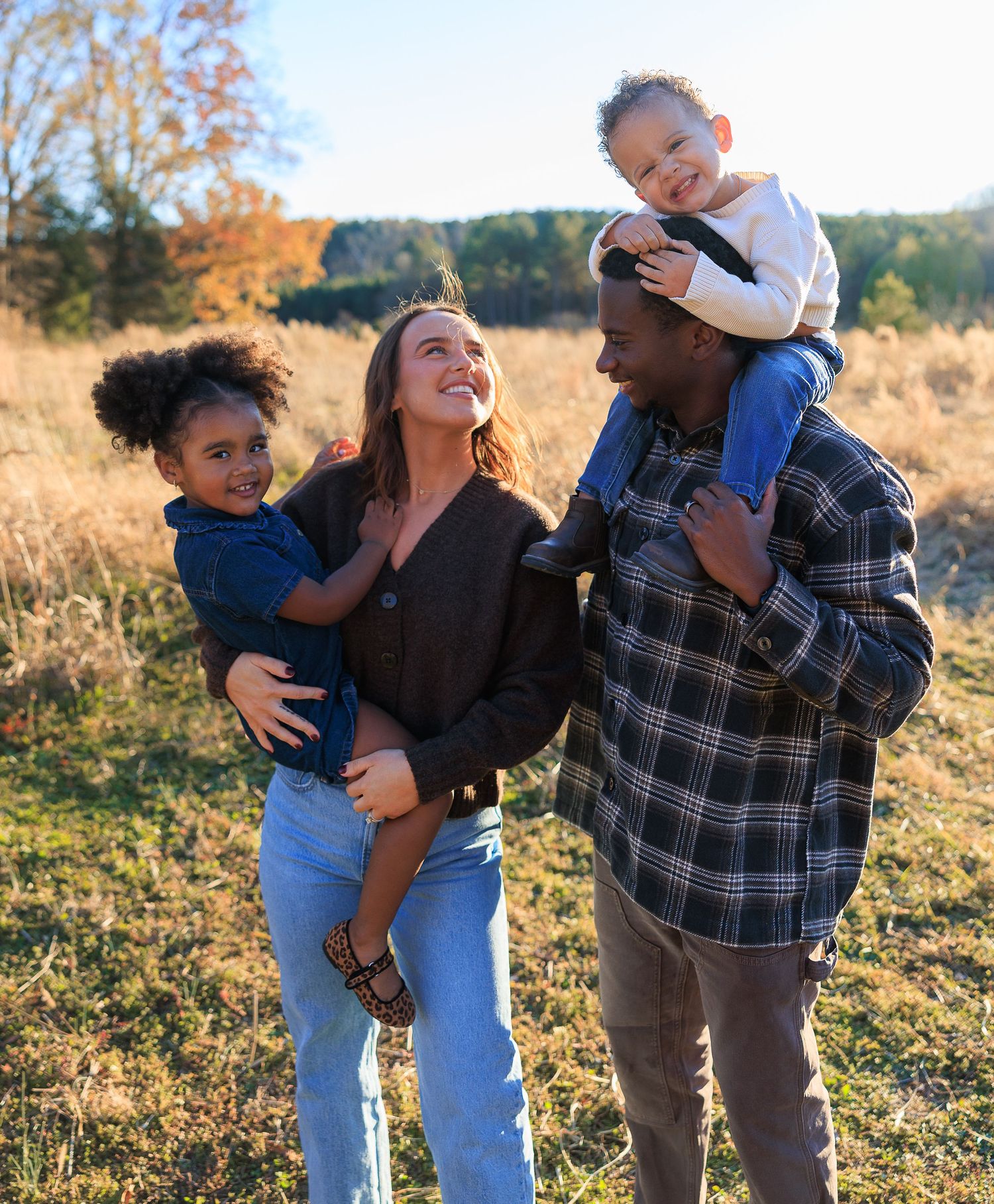 A family enjoys a warm autumn day together in a sunlit field with golden grass and fall foliage in the background.