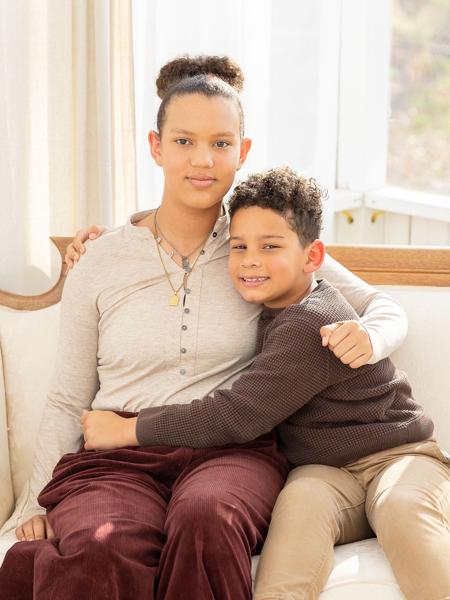 Two siblings hugging on a white sofa, older teen in gray top with younger child in brown sweater.