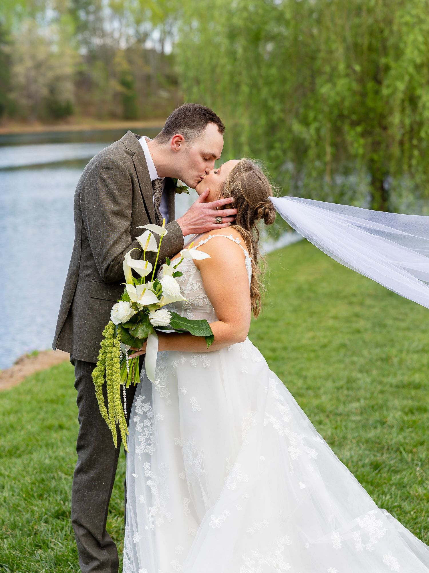 Bride and groom share romantic kiss outdoors, veil flowing in breeze, holding white calla lily bouquet near serene pond.