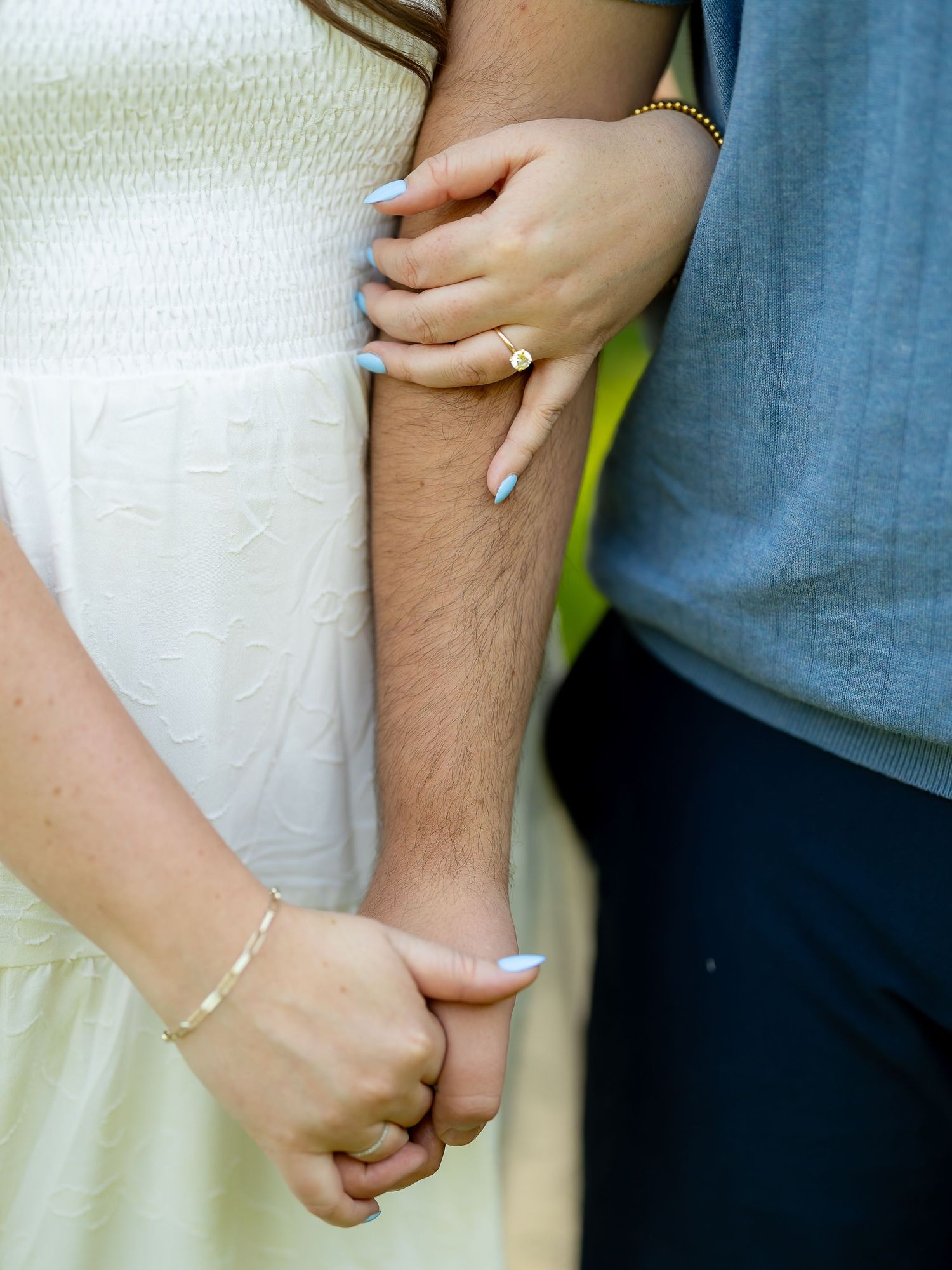 Close-up of engaged couple holding hands, woman wearing gold ring and bracelet with light blue nails.