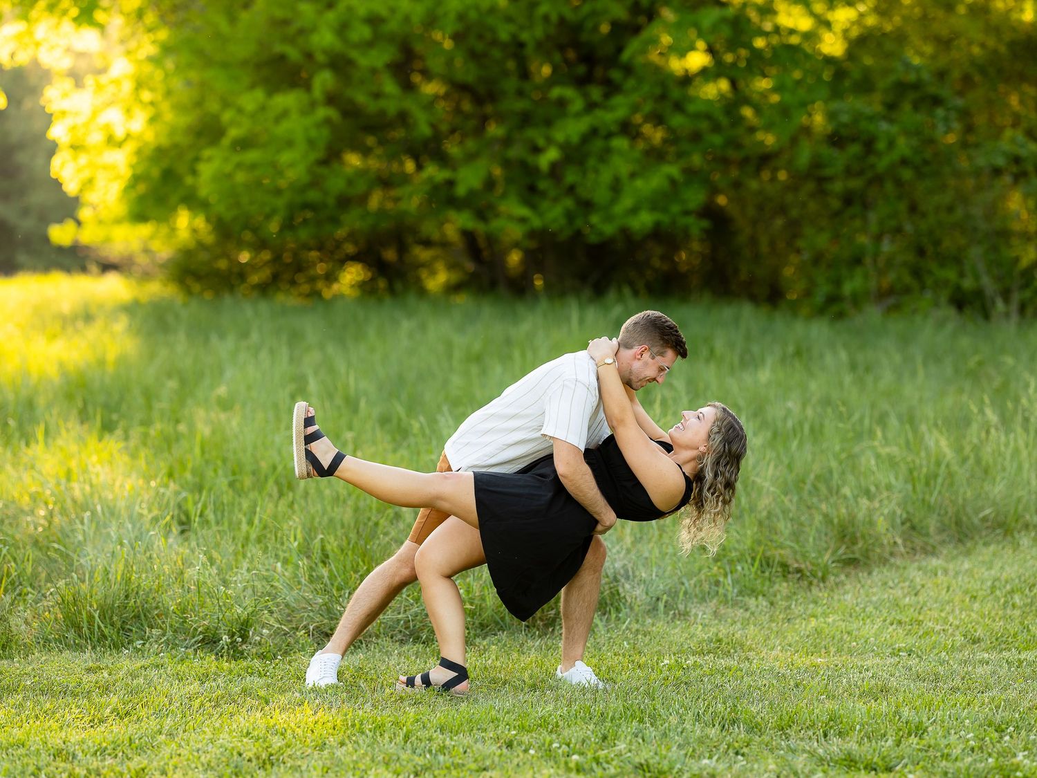 Couple sharing a romantic dip kiss in a lush green meadow during golden hour outdoor engagement photo.