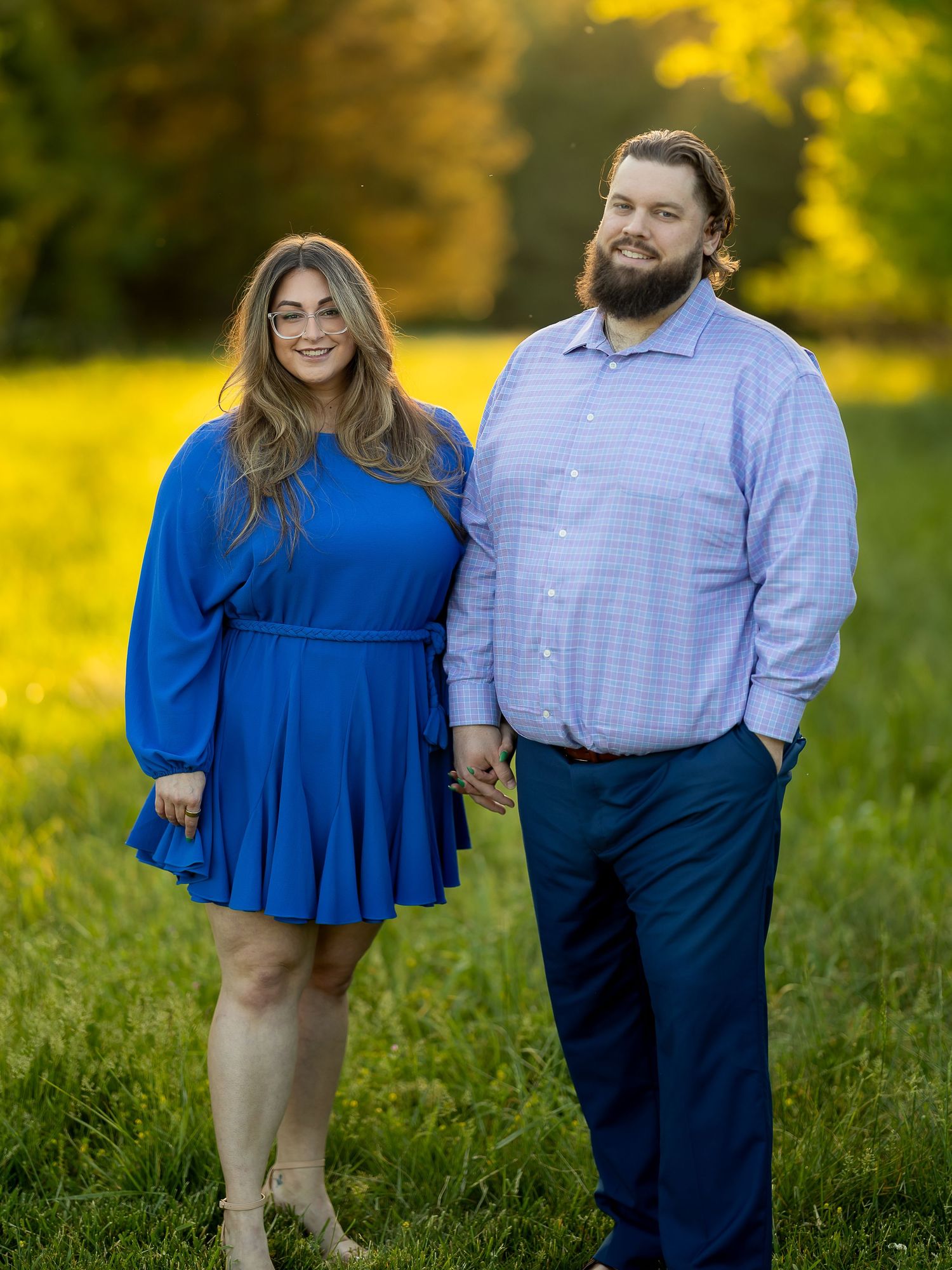 Couple holding hands outdoors in a sunny field, woman in blue dress and man in blue shirt and navy pants.