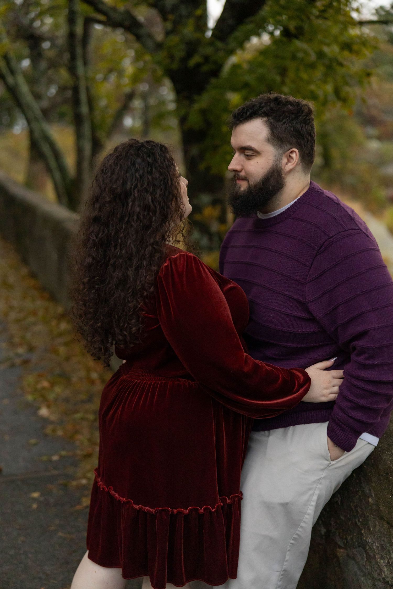A couple in burgundy velvet and purple sweater embraces in an autumn park setting with fallen leaves.