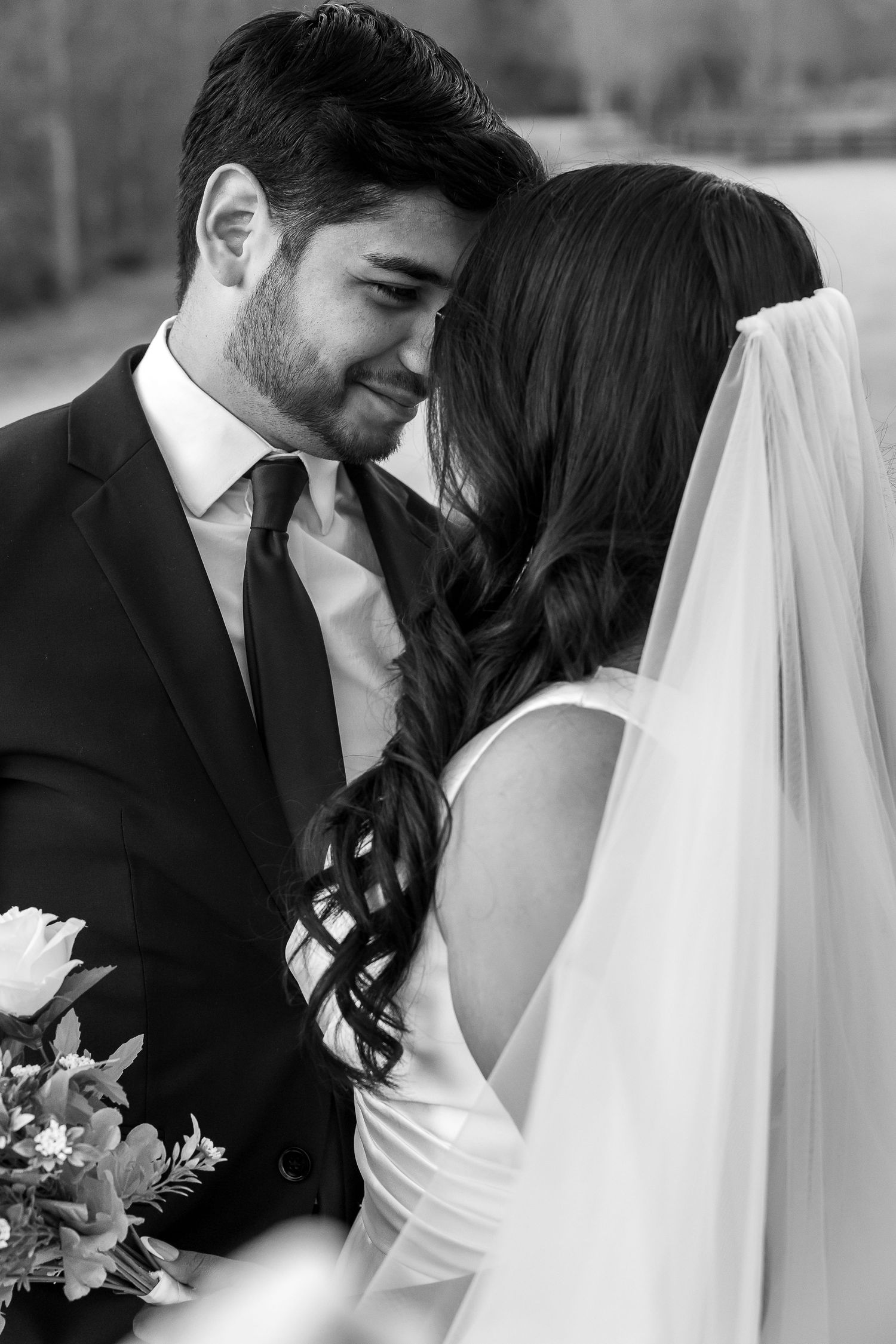 Black and white close-up of groom gazing at bride in wedding veil, holding floral bouquet.