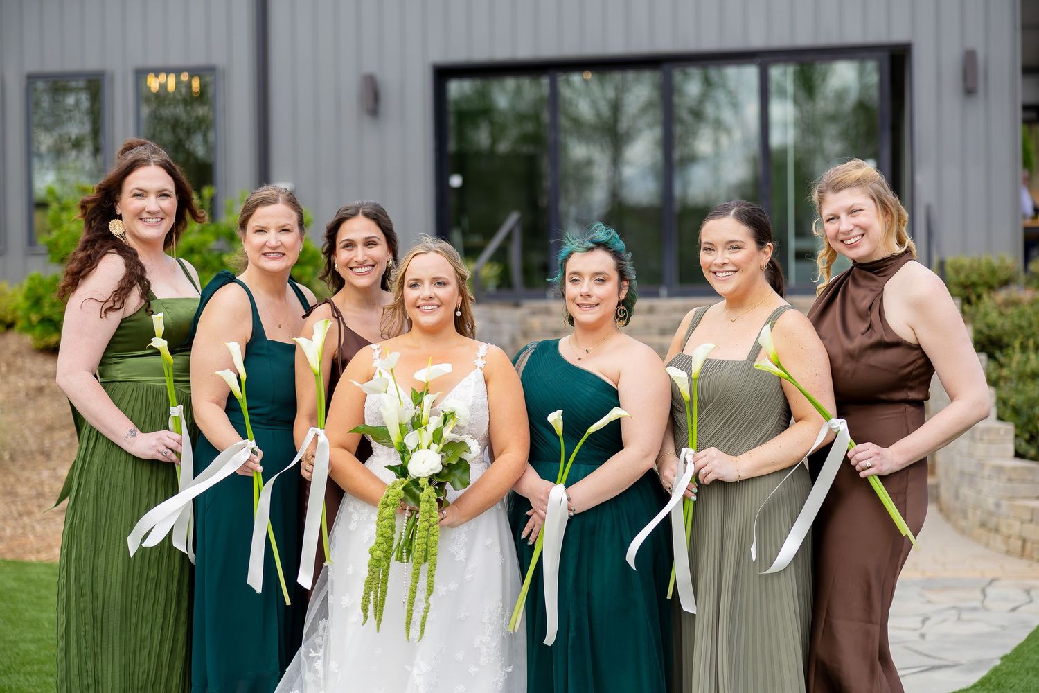 Bride poses with six bridesmaids in green and brown dresses holding white ribbon bouquets outside a modern venue.