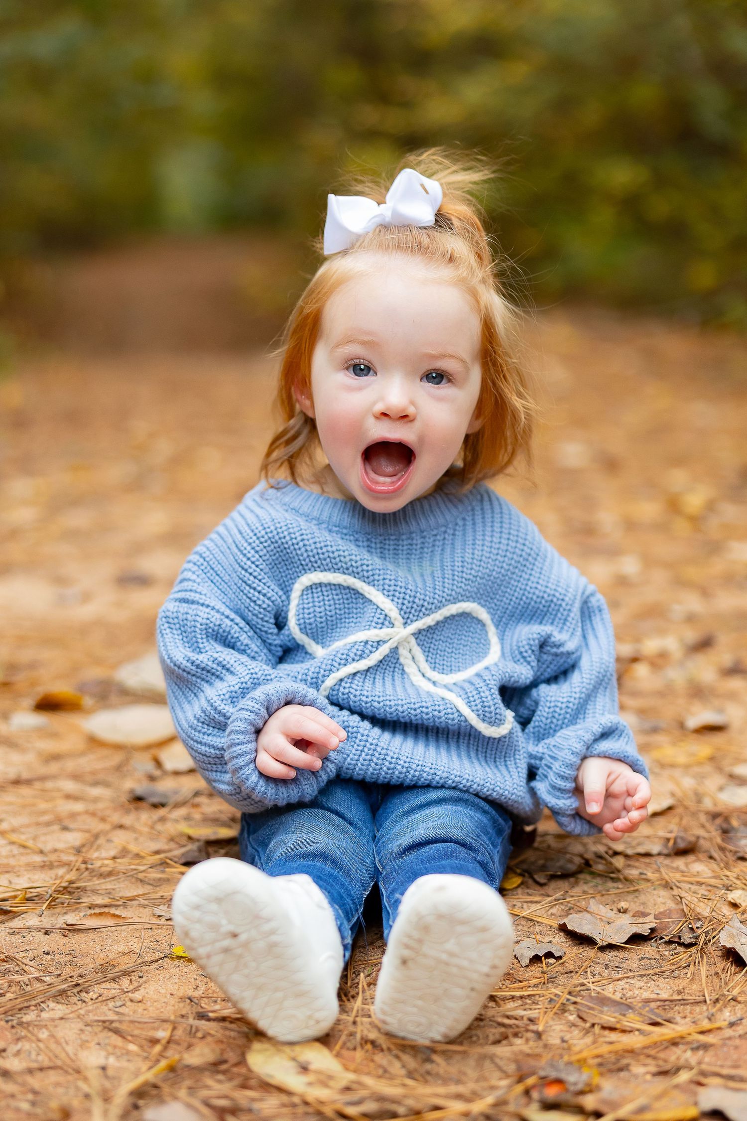 A baby in a blue sweater sits on fallen autumn leaves with an excited expression.