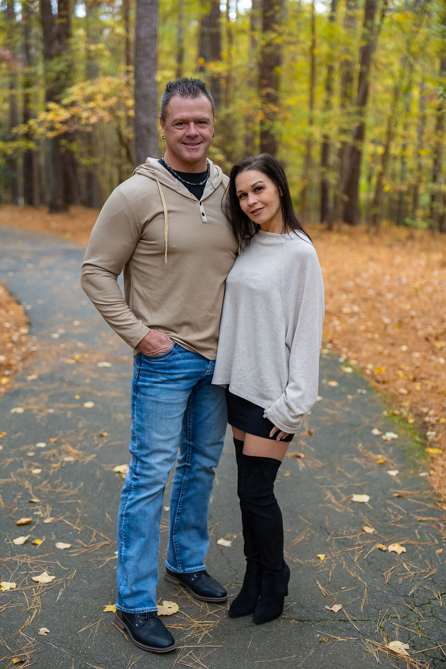 A couple poses together on a path surrounded by autumn trees with golden leaves scattered on the ground.