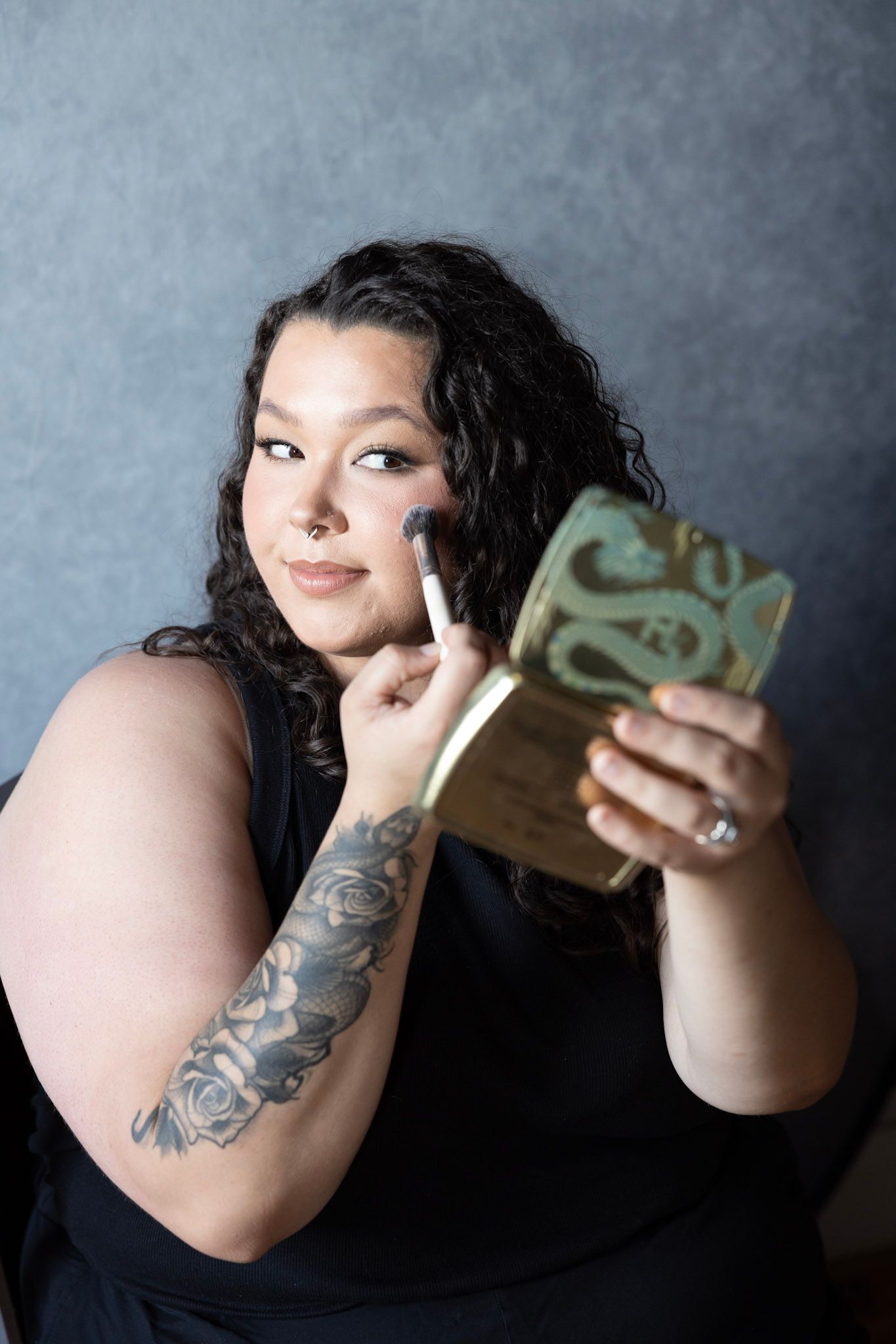 Person in black showing tattoo art while holding up items against grey backdrop.