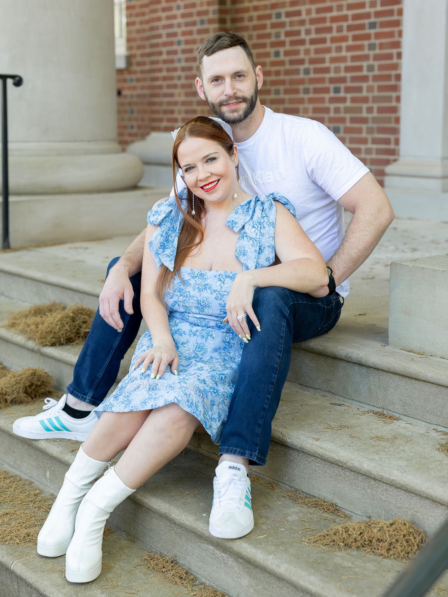 A smiling couple sitting together on outdoor stairs, the woman wearing a blue floral dress and the man wearing a white shirt.