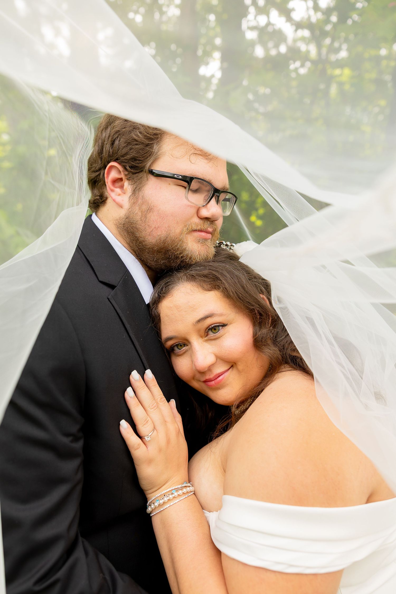 Bride and groom embrace tenderly under a flowing white veil outdoors on their wedding day.