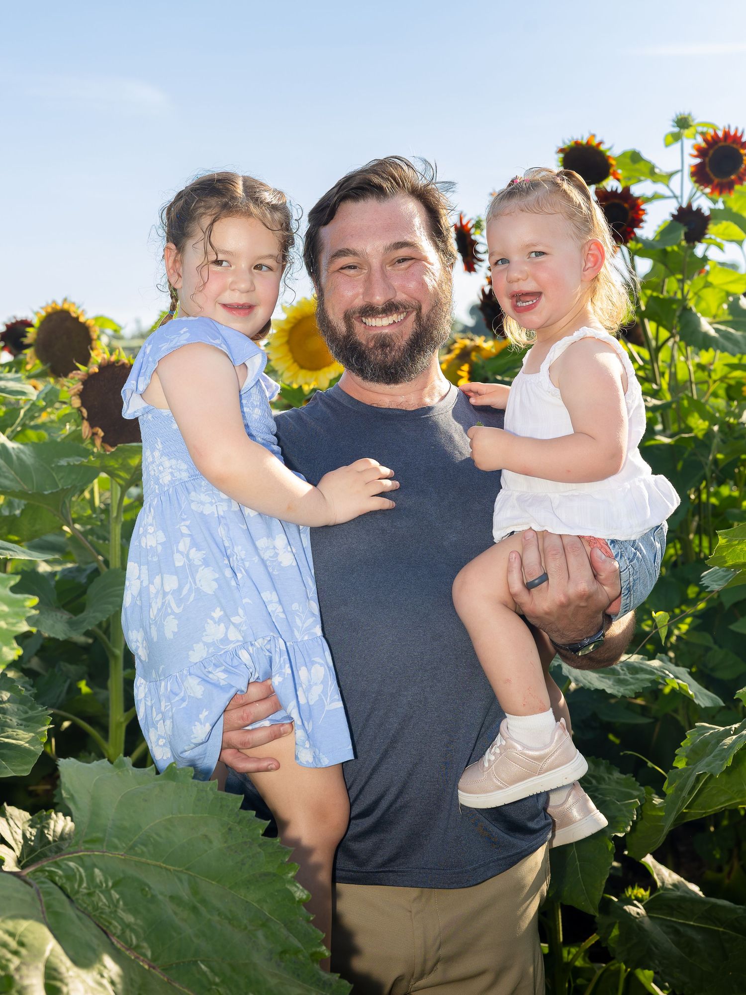 A smiling bearded man holds two young girls in a sunny sunflower field, all dressed casually and enjoying the outdoors.