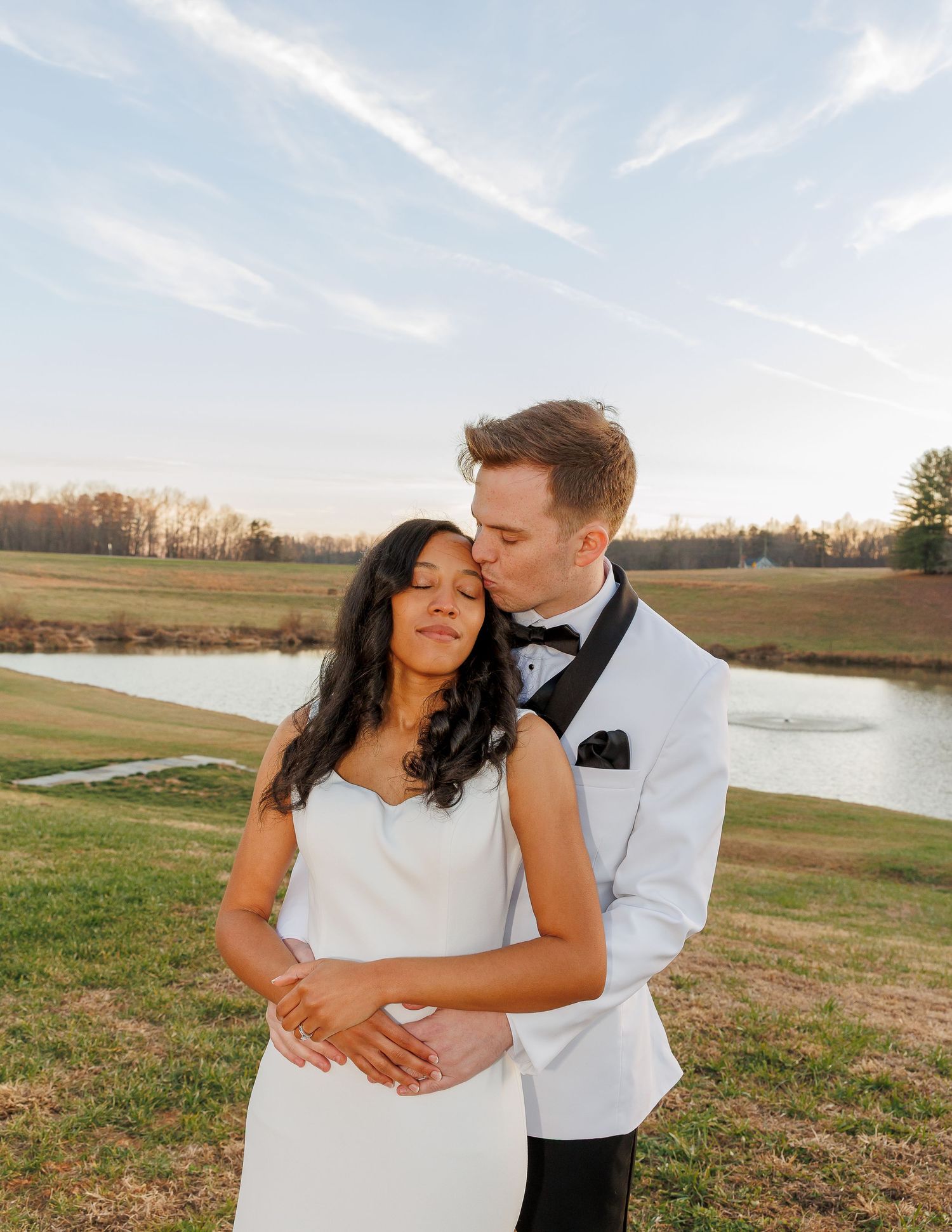 Groom in white tuxedo embraces bride in white dress outdoors near pond at golden hour wedding.