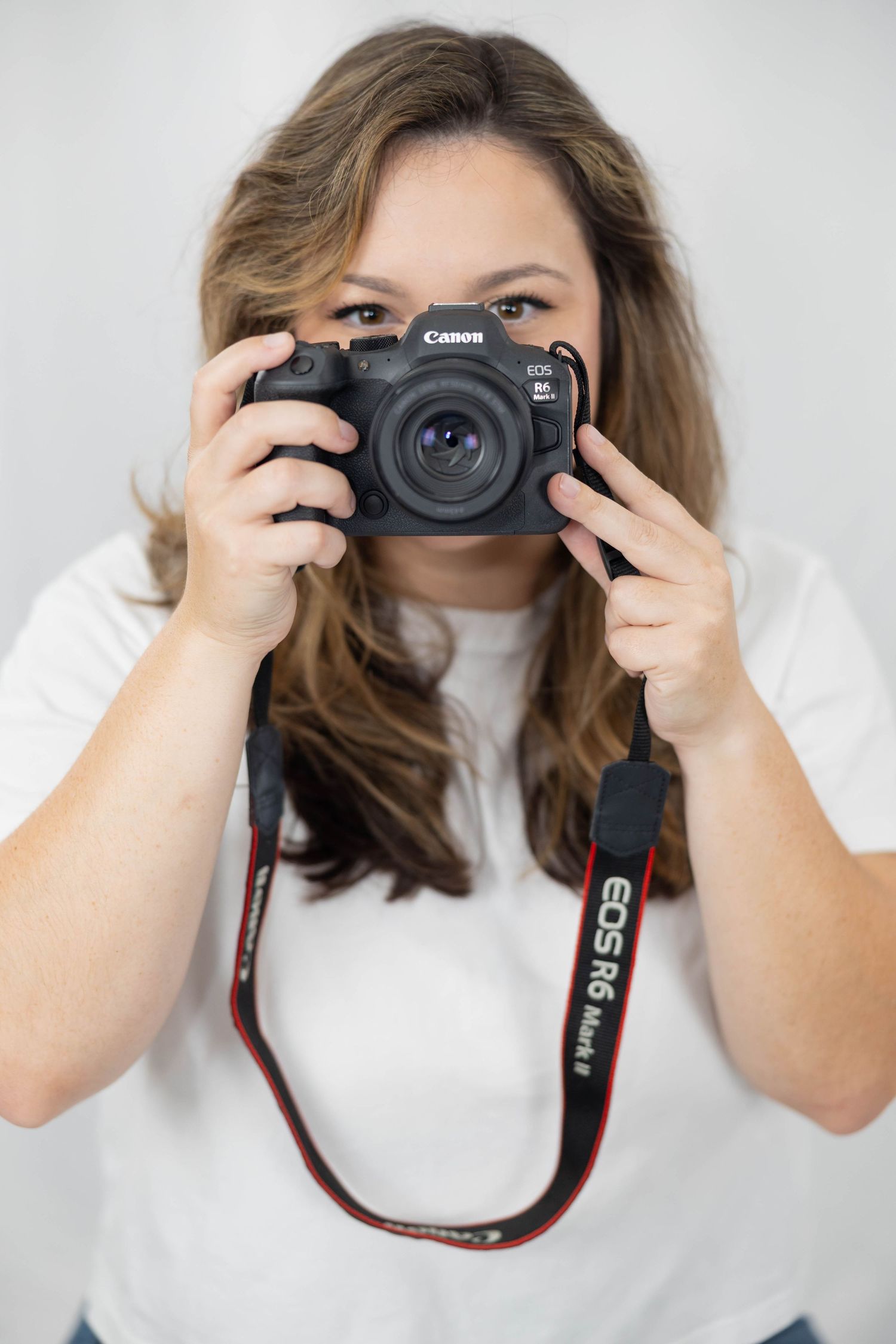 A photographer wearing a white t-shirt takes a photo with a Canon DSLR camera and red camera strap.