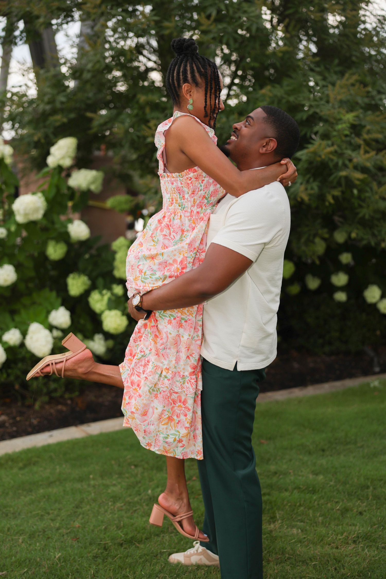 Romantic couple embraces in a garden setting with white roses, the woman wearing a floral pink dress lifted up by her partner.