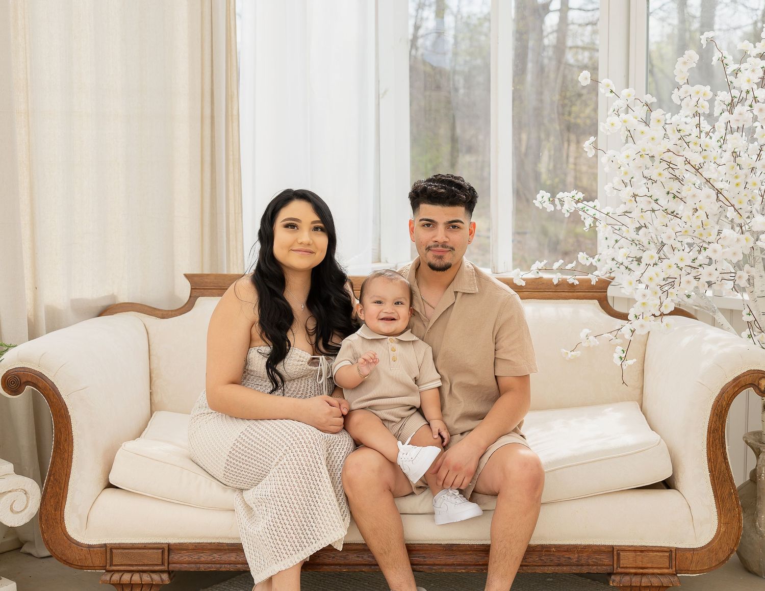A family lounges together on a cream-colored sofa in a bright, sunlit living room with white decor.