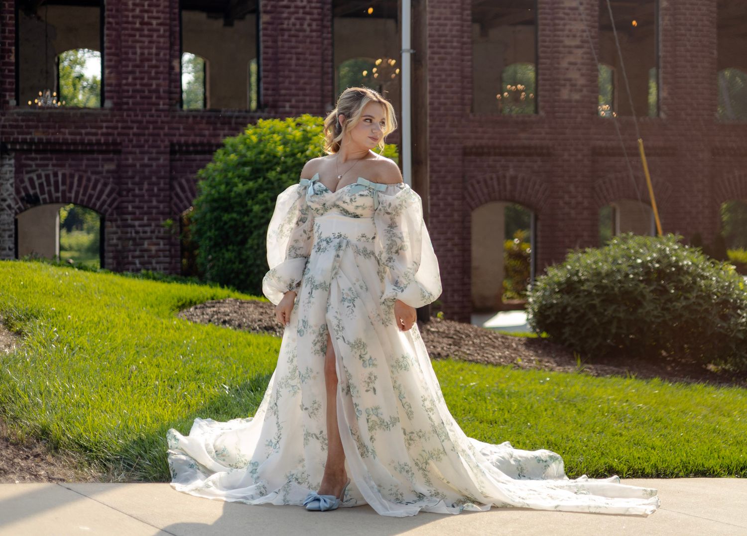 A bride in a floral lace wedding dress poses for a series of outdoor portraits in front of a brick building.