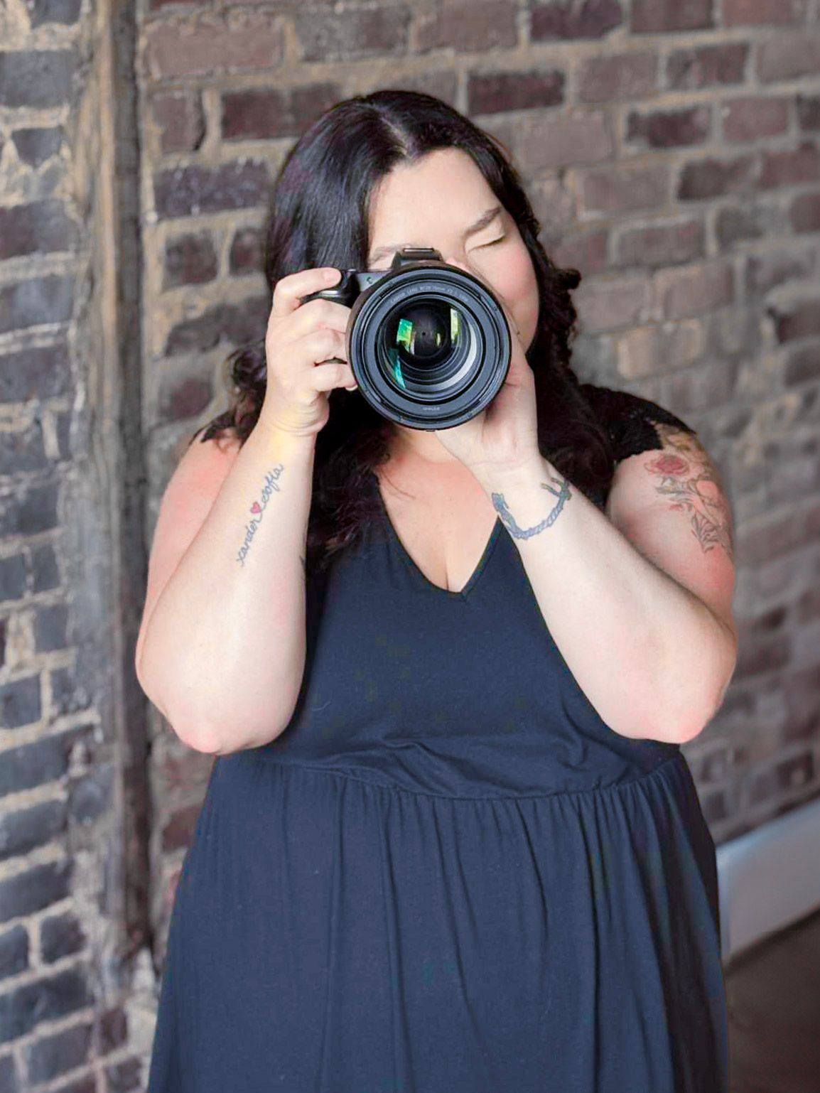 Professional photographer in a navy dress holding a camera with large lens against brick wall.
