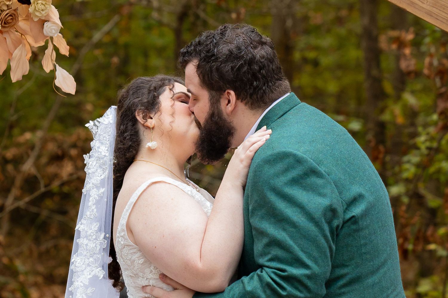 Bride and groom share first kiss at outdoor fall wedding ceremony with autumn foliage backdrop.