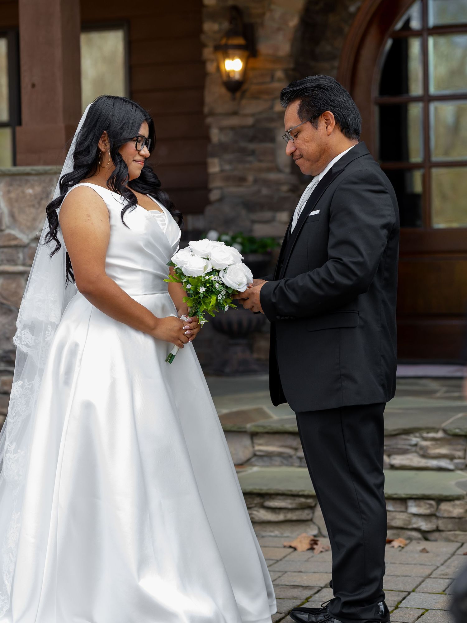 Bride in white gown holding bouquet and groom in black tuxedo share a tender moment outside a stone building.