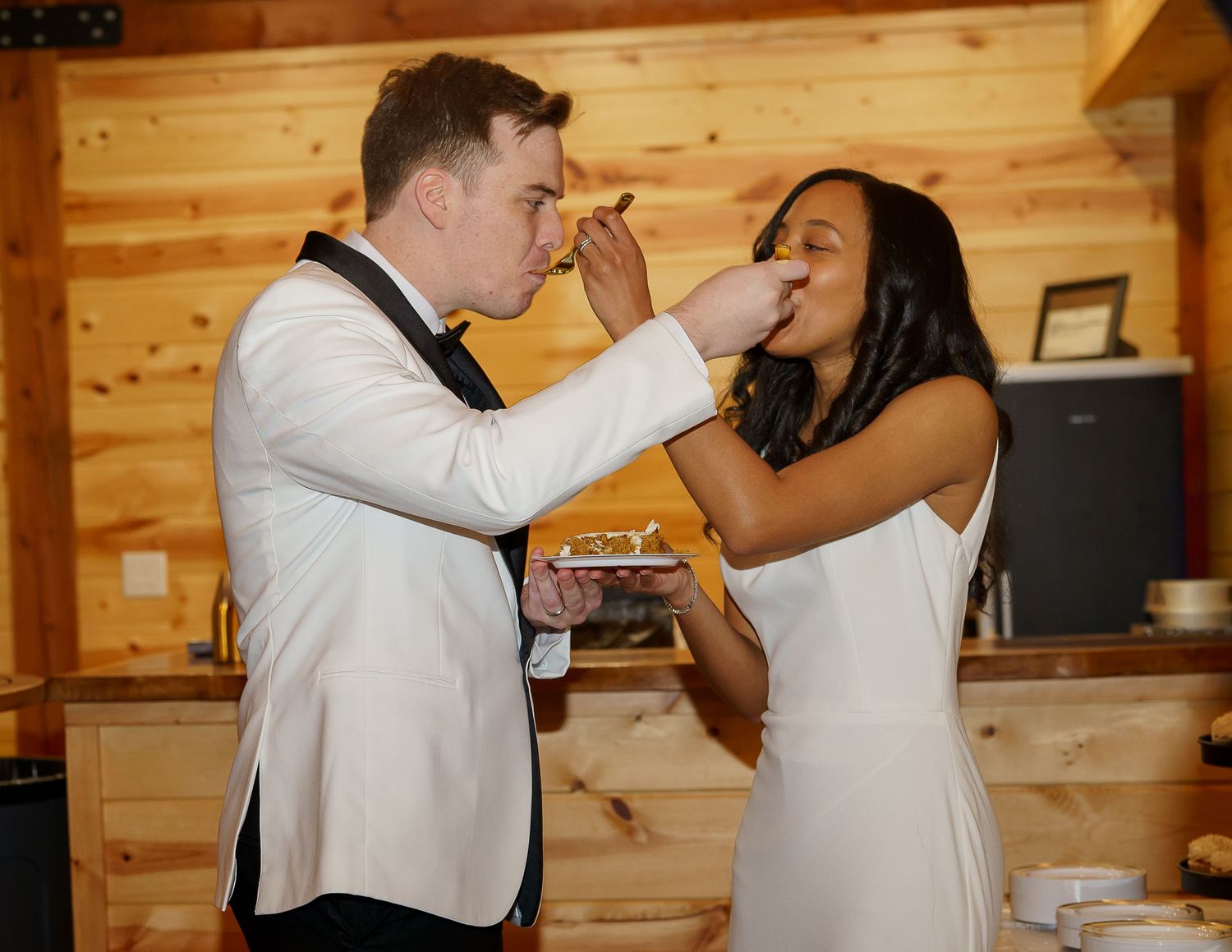 Newlyweds playfully feed each other wedding cake inside a rustic wooden venue during their reception.