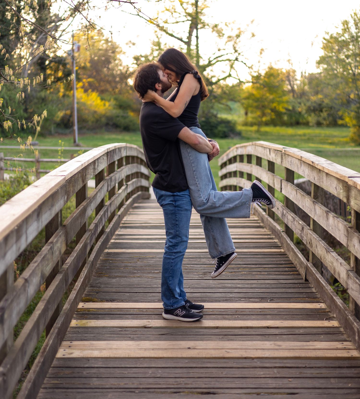 Romantic couple embracing on a wooden bridge in a park during golden hour sunset lighting.