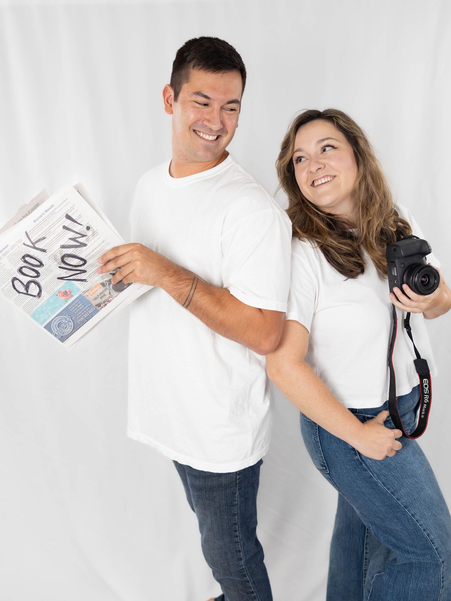 Two people in white t-shirts and jeans sharing a moment with a book and camera against a light background.