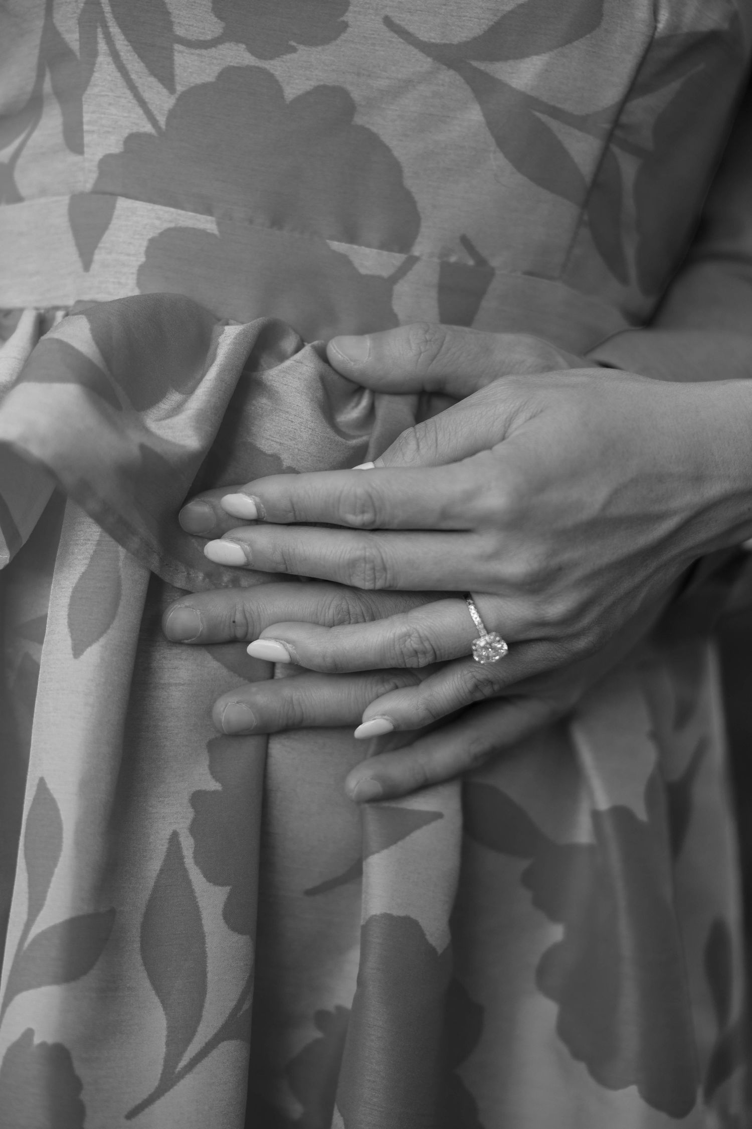 Black and white image of hands showing off an engagement ring against a floral patterned fabric.