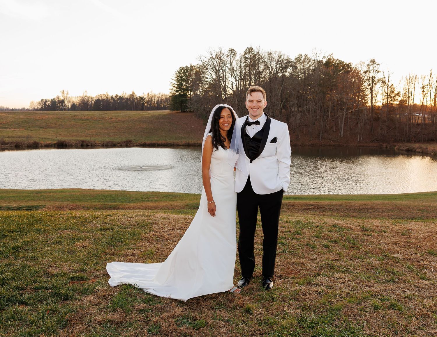 A bride and groom stand together on a grassy hill at sunset with a serene pond in the background.