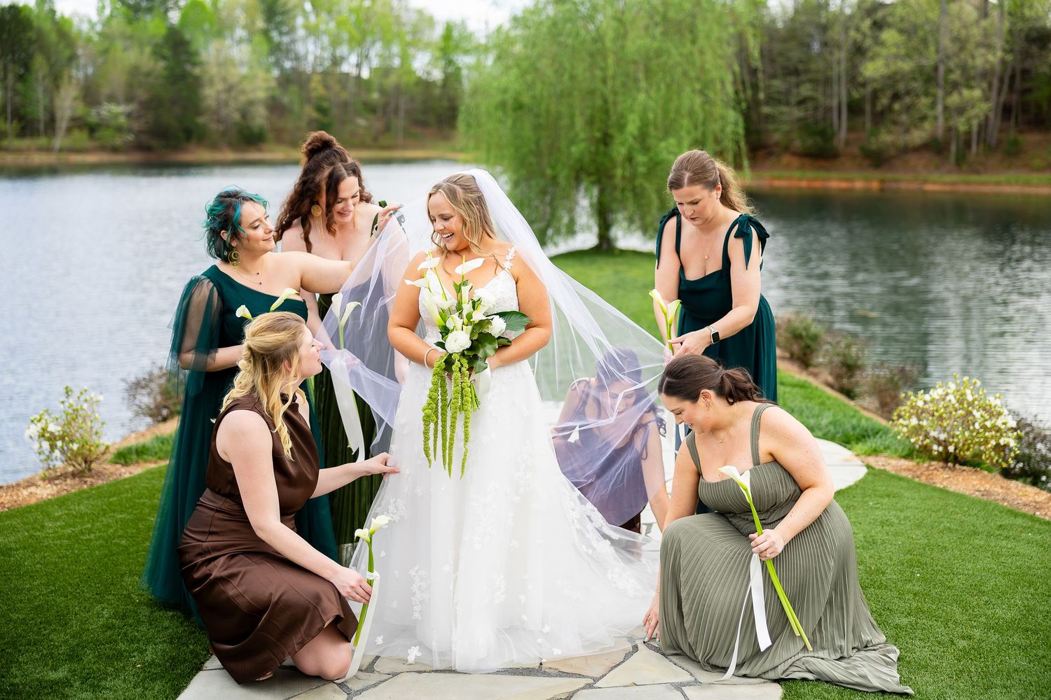 Bride in white gown with veil surrounded by bridesmaids in green dresses near a lake outdoors.