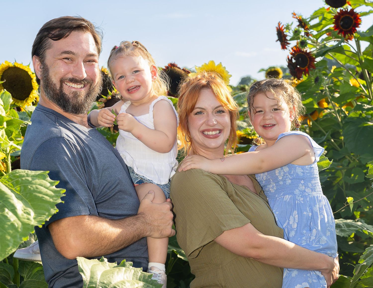 Happy family of four posing in a sunflower field on a sunny day, with two young children and smiling parents.