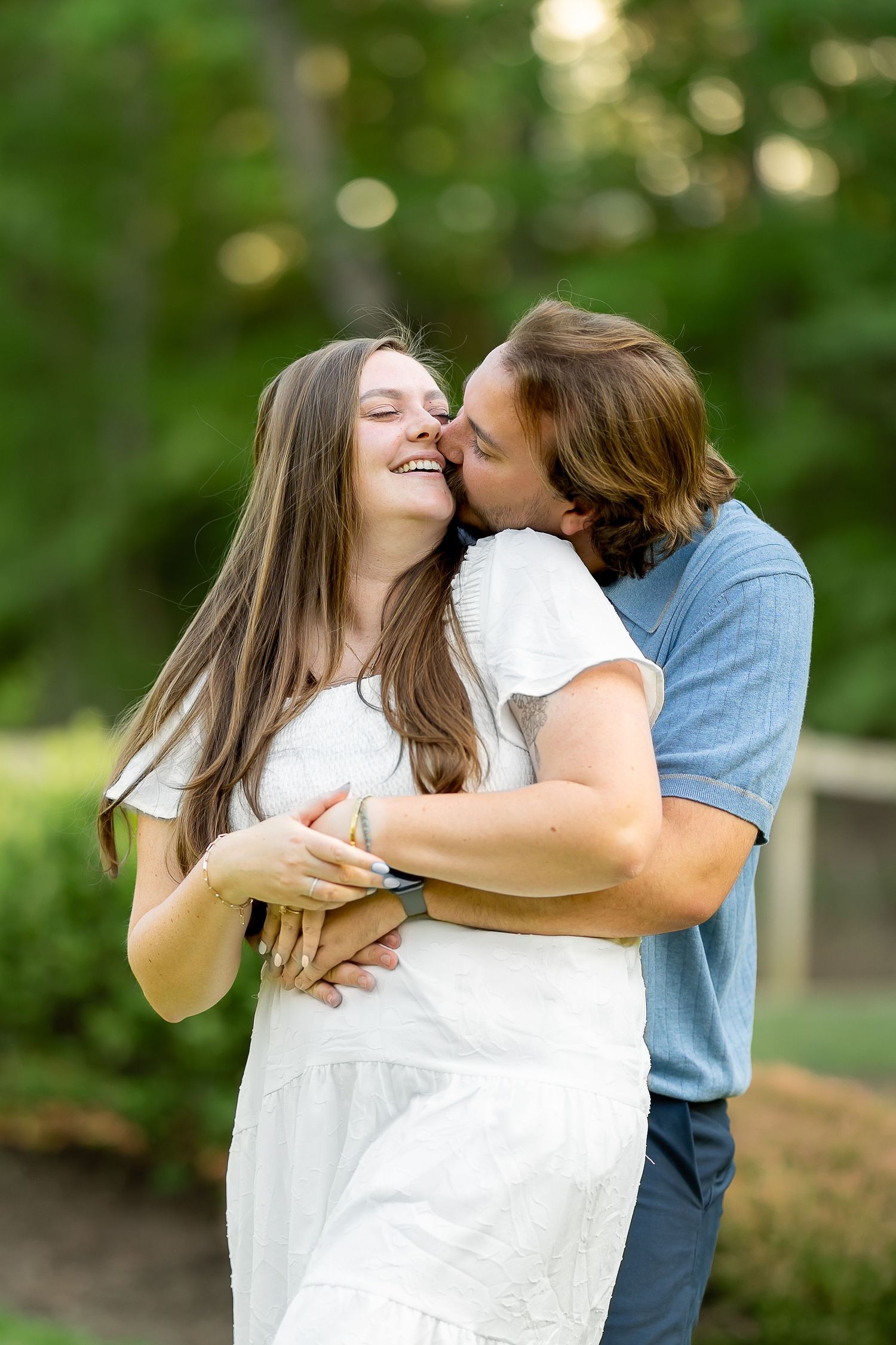 A couple embraces outdoors, man kissing woman's cheek as she laughs, surrounded by lush green trees.