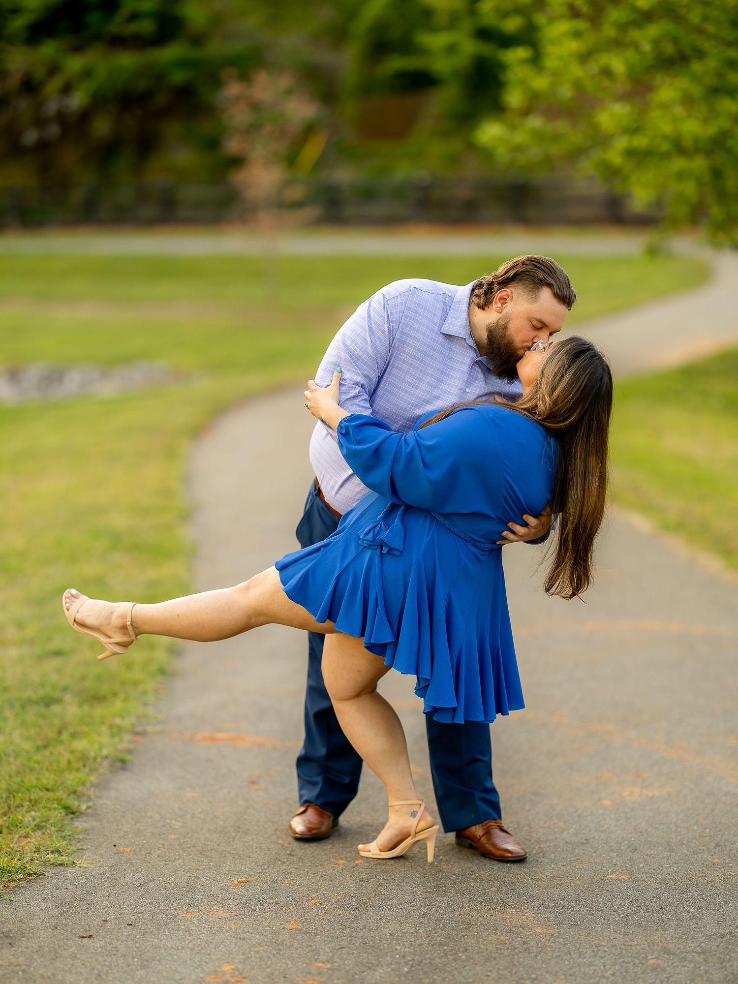 Couple sharing a romantic dip kiss on a park path, woman in blue dress and heels, man in blue shirt and dark pants.