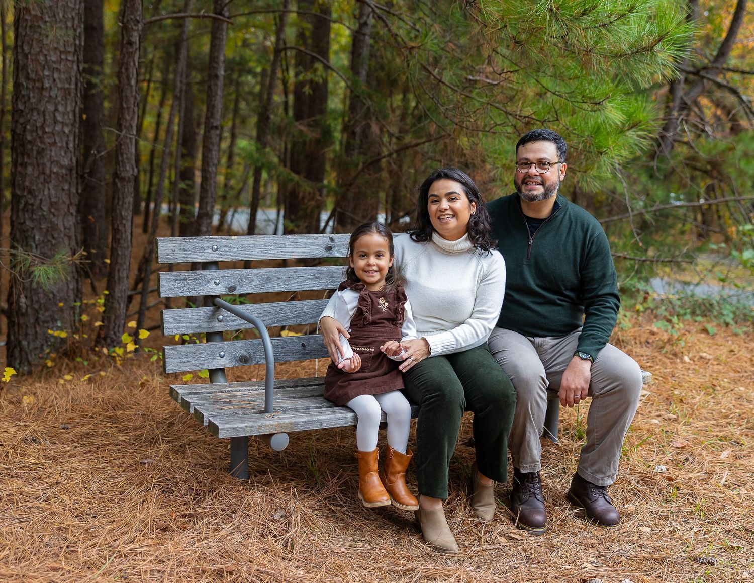 A family sits together on a wooden bench along a peaceful forest trail covered in pine needles.
