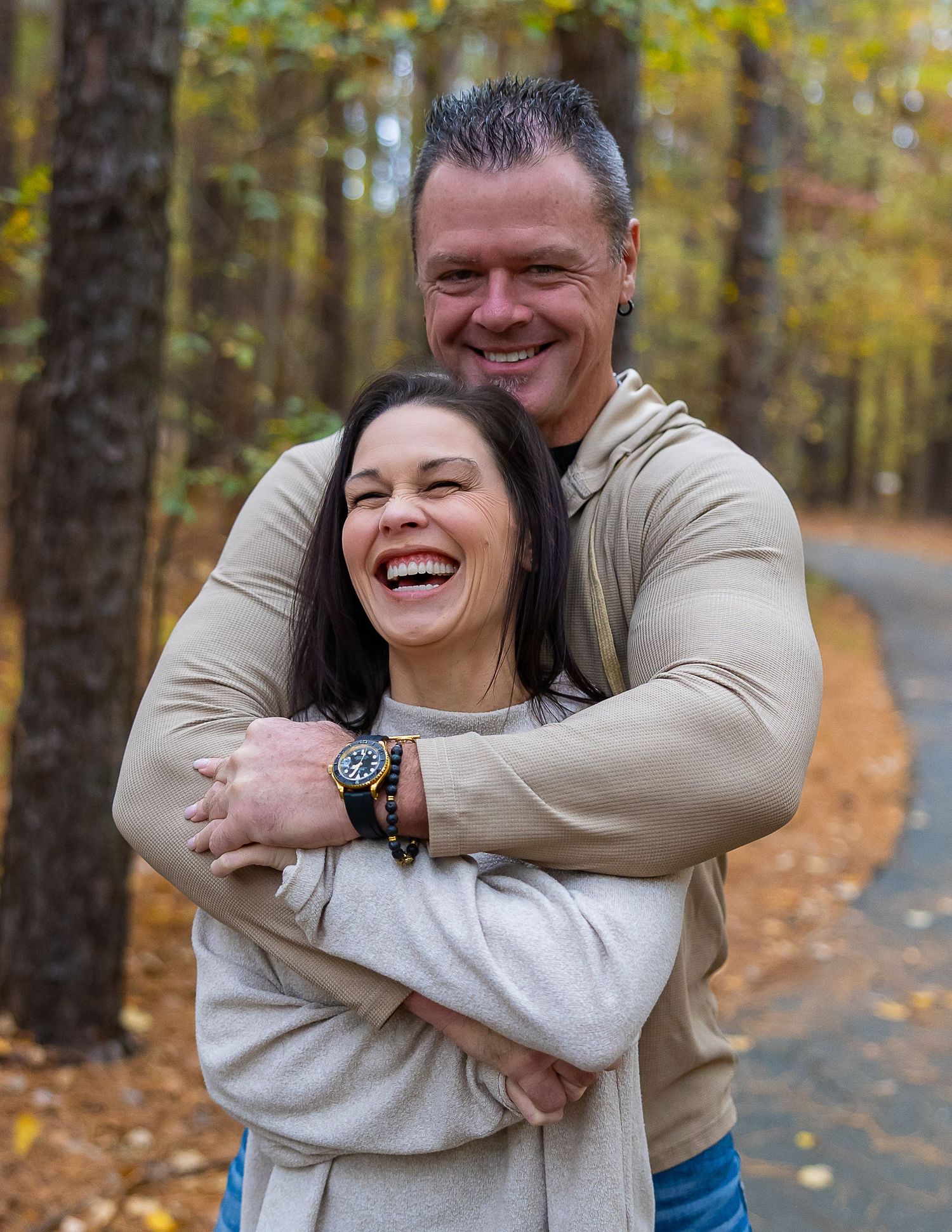 A couple shares a joyful embrace while standing on a path surrounded by autumn trees with golden foliage.