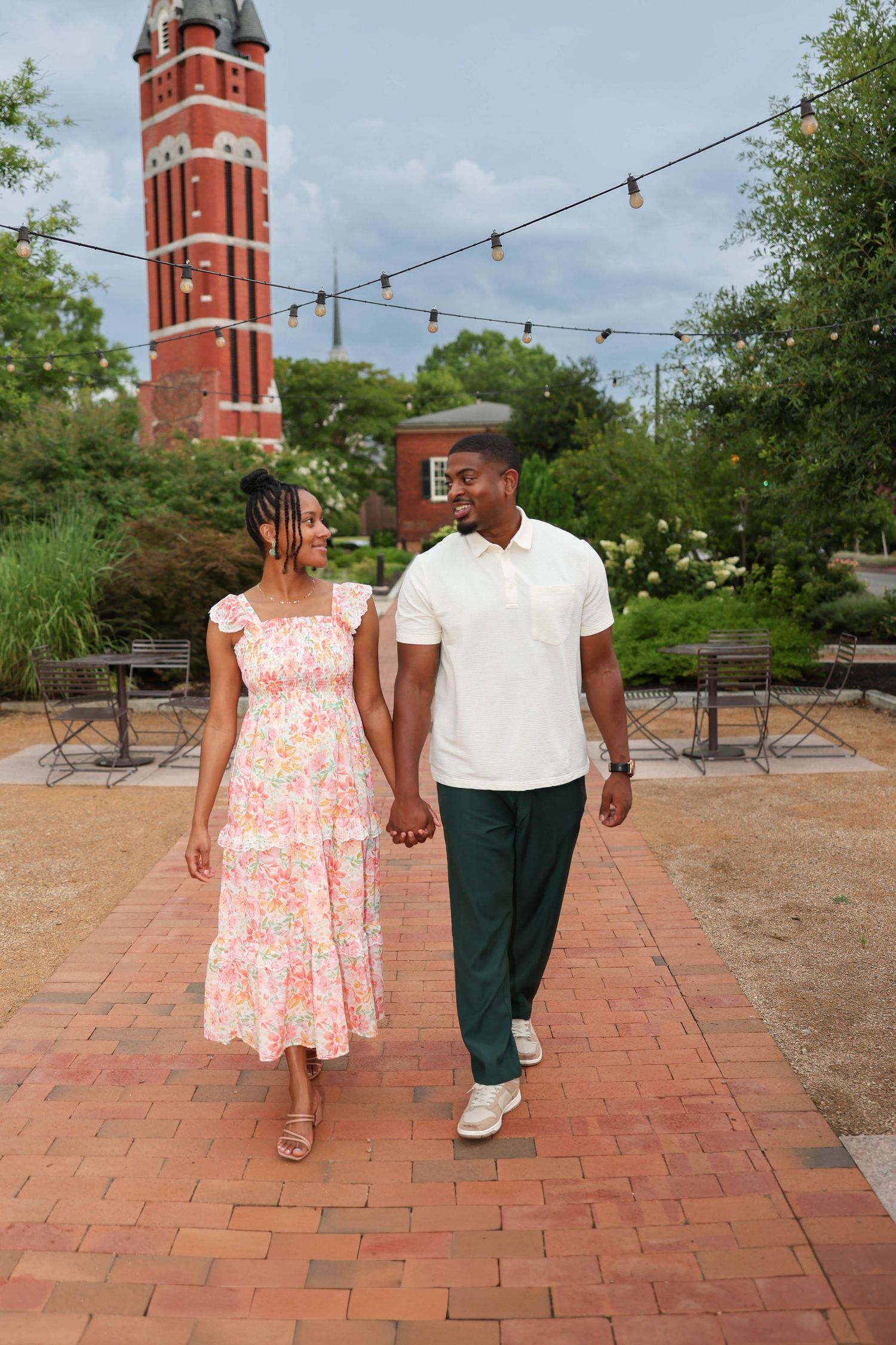 A couple strolls hand in hand on brick pavement with a red brick bell tower and string lights visible in the background.