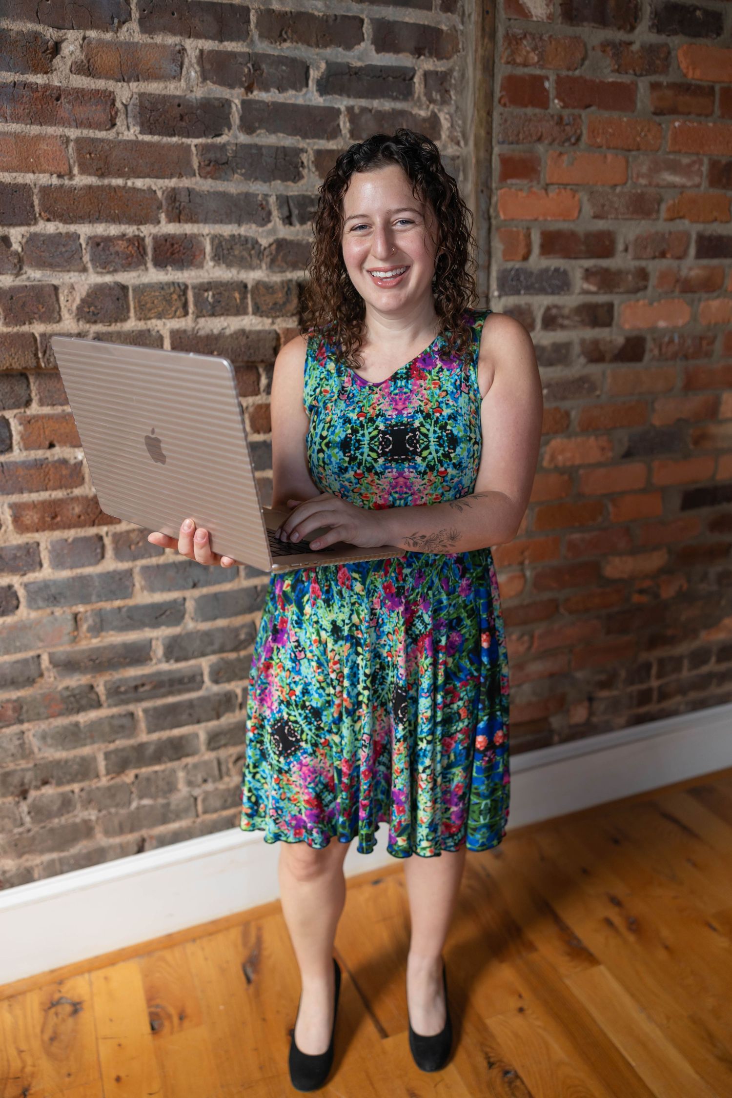 Person in a colorful floral dress stands against exposed brick wall while holding a MacBook laptop.