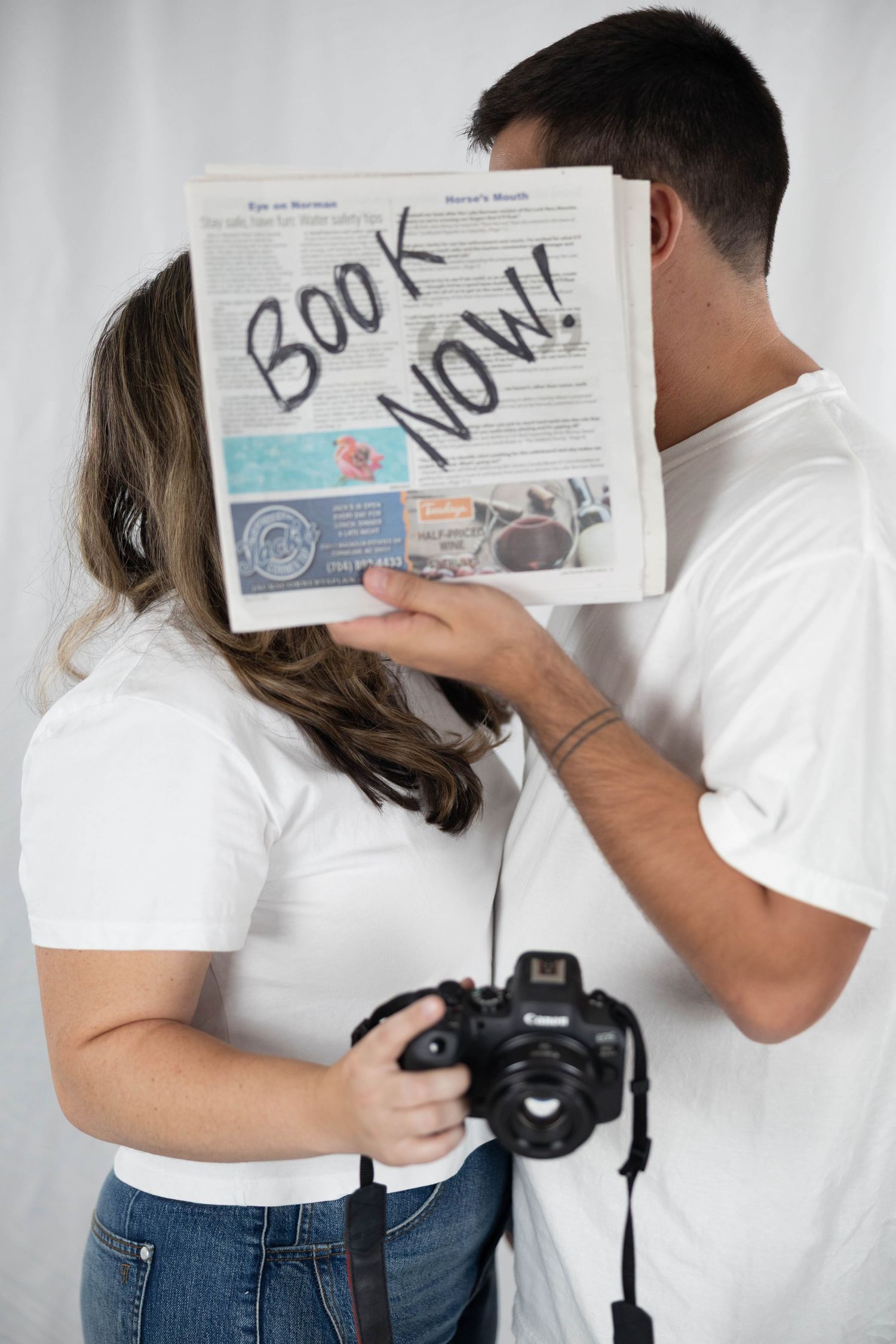 Couple poses together while holding and reading an information card and holding a camera against white background.