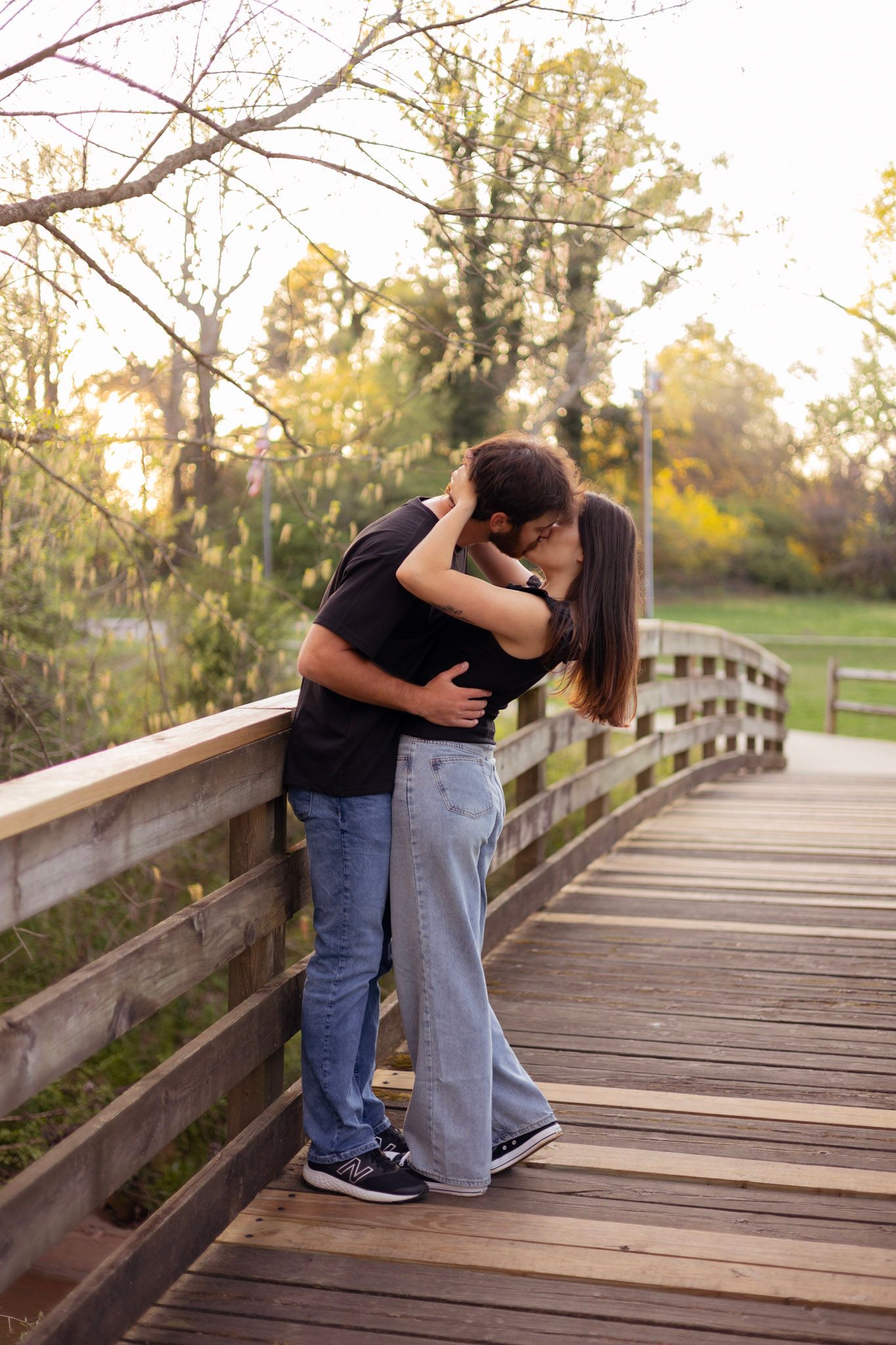 A couple shares an intimate kiss while embracing on a wooden bridge walkway surrounded by autumn trees at sunset.