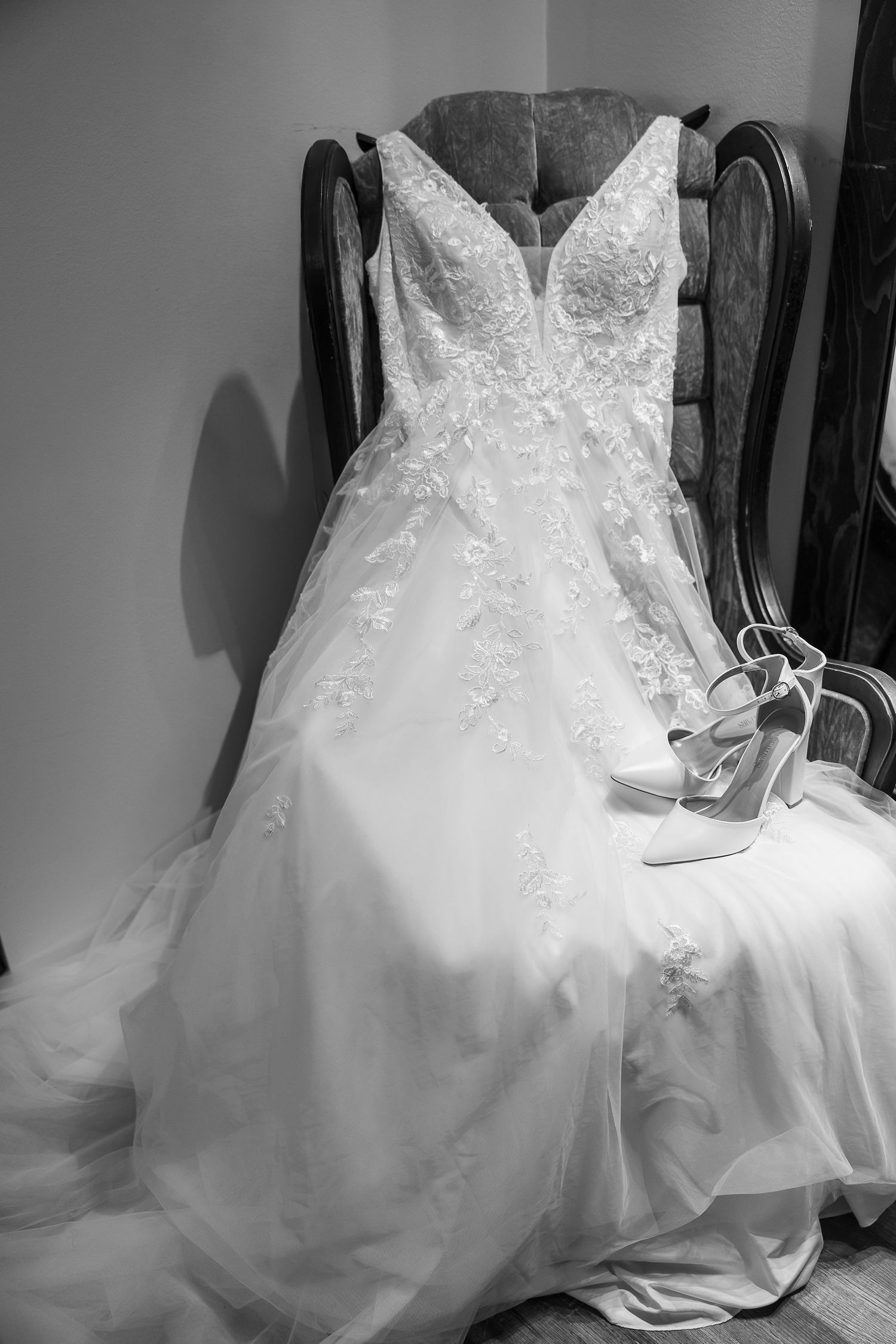 Black and white photo of an elegant lace wedding gown draped over a chair with white heeled bridal shoes.