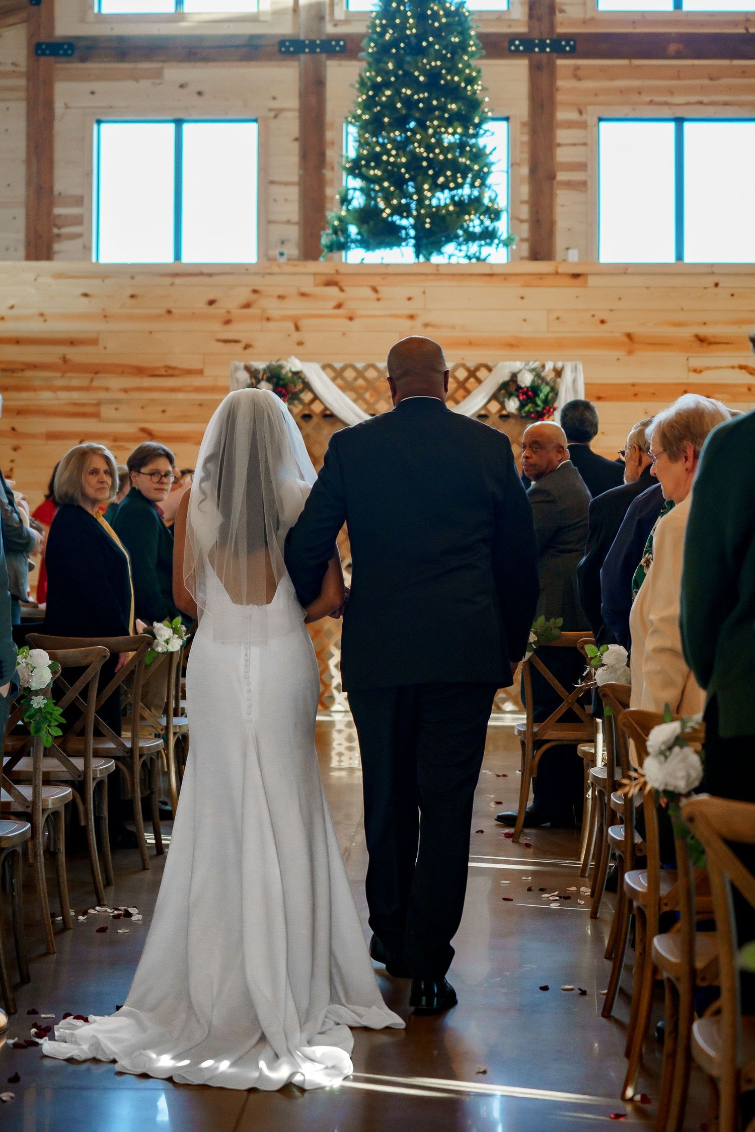 Bride walking down the aisle with escort in rustic barn wedding venue with Christmas tree.