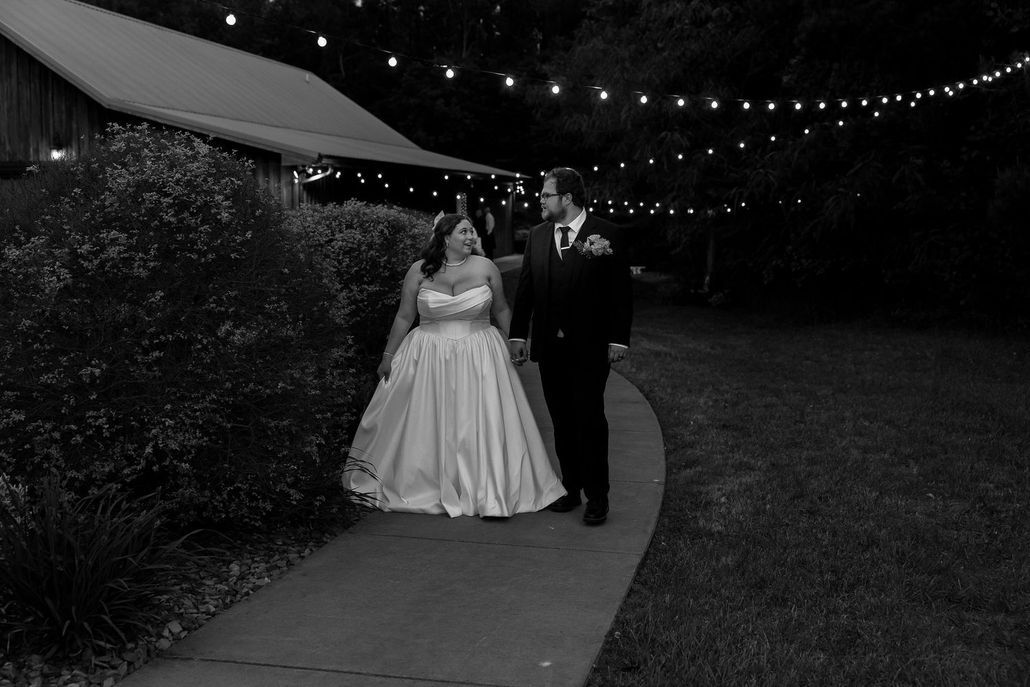 Bride and groom walk together under string lights in a romantic black and white wedding photo.