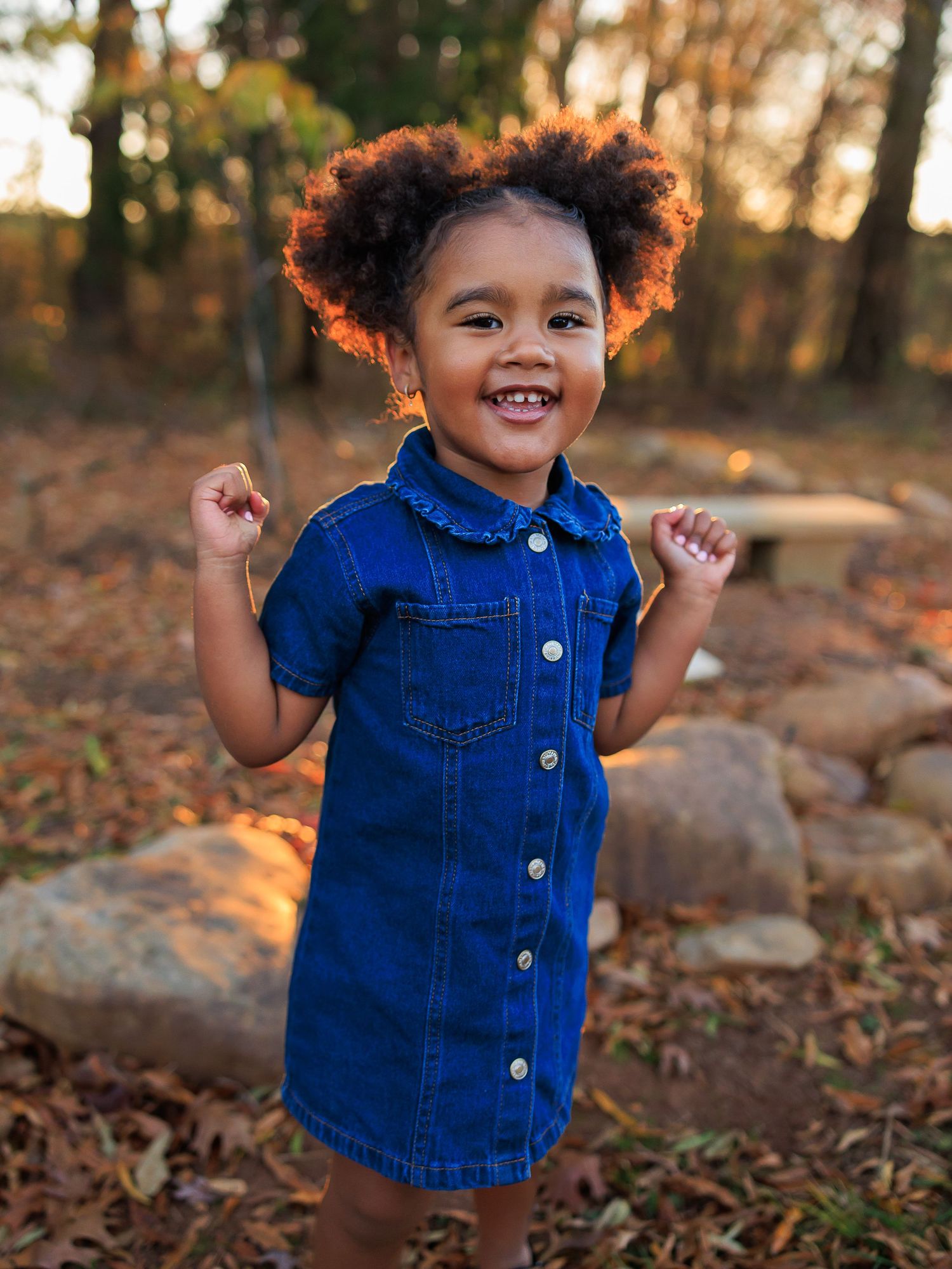 A young child in a denim button-up dress smiles joyfully while standing outdoors among autumn leaves and rocks.