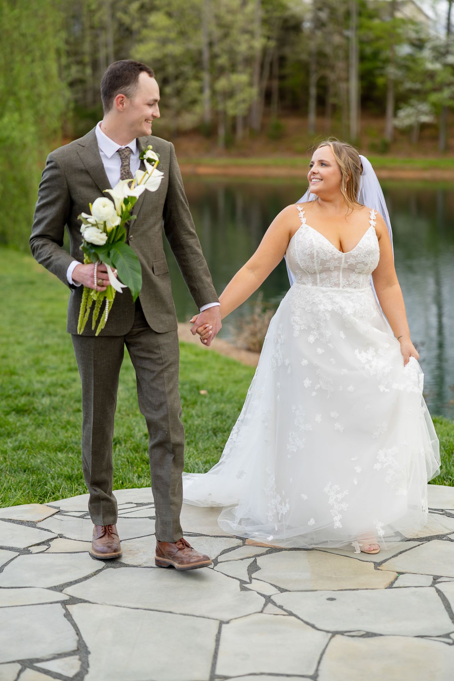 A smiling bride and groom hold hands while walking on stone pavers near a pond, she in a white gown, he in a gray suit.
